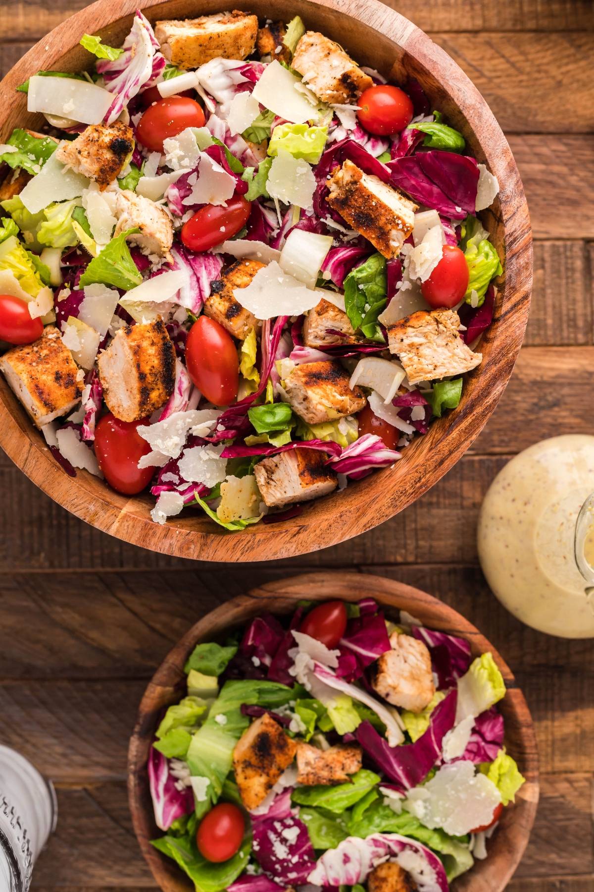 Two wooden bowls of grilled chicken Caesar salad with cherry tomatoes and shaved parmesan, reminiscent of a summer BBQ chicken salad, sit on a wooden table.