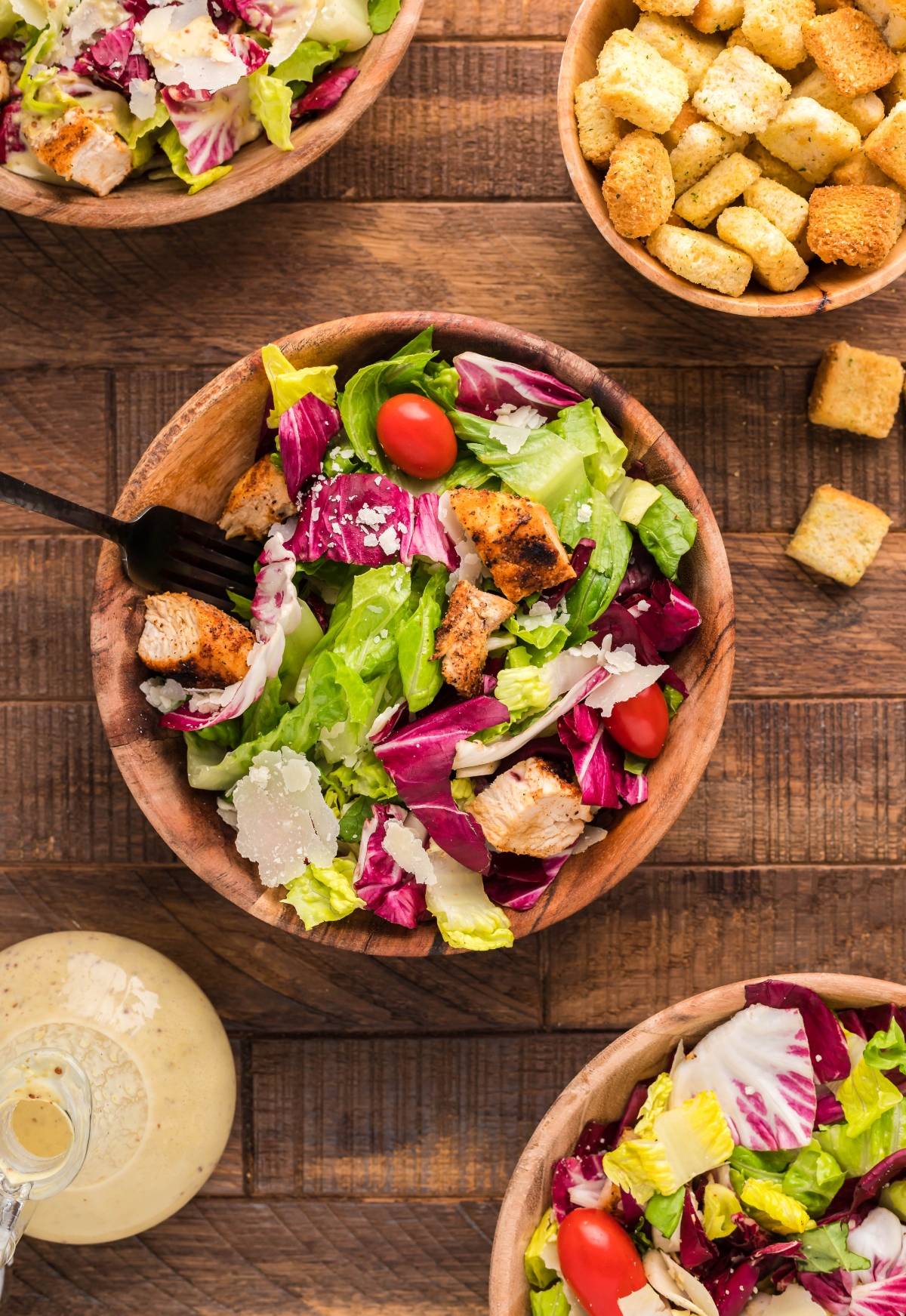 Wooden bowls of summer BBQ chicken salad with lettuce, cherry tomatoes, grilled chicken, croutons, and shaved parmesan on a table.