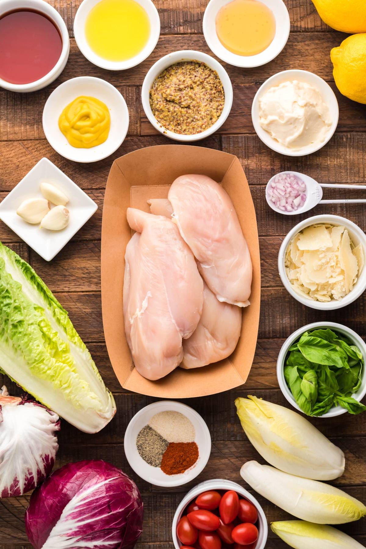 Raw chicken breasts and various fresh salad ingredients, including endive and radicchio for a simple radicchio salad, are arranged on a wooden table—perfect for crafting a vibrant summer BBQ chicken salad.