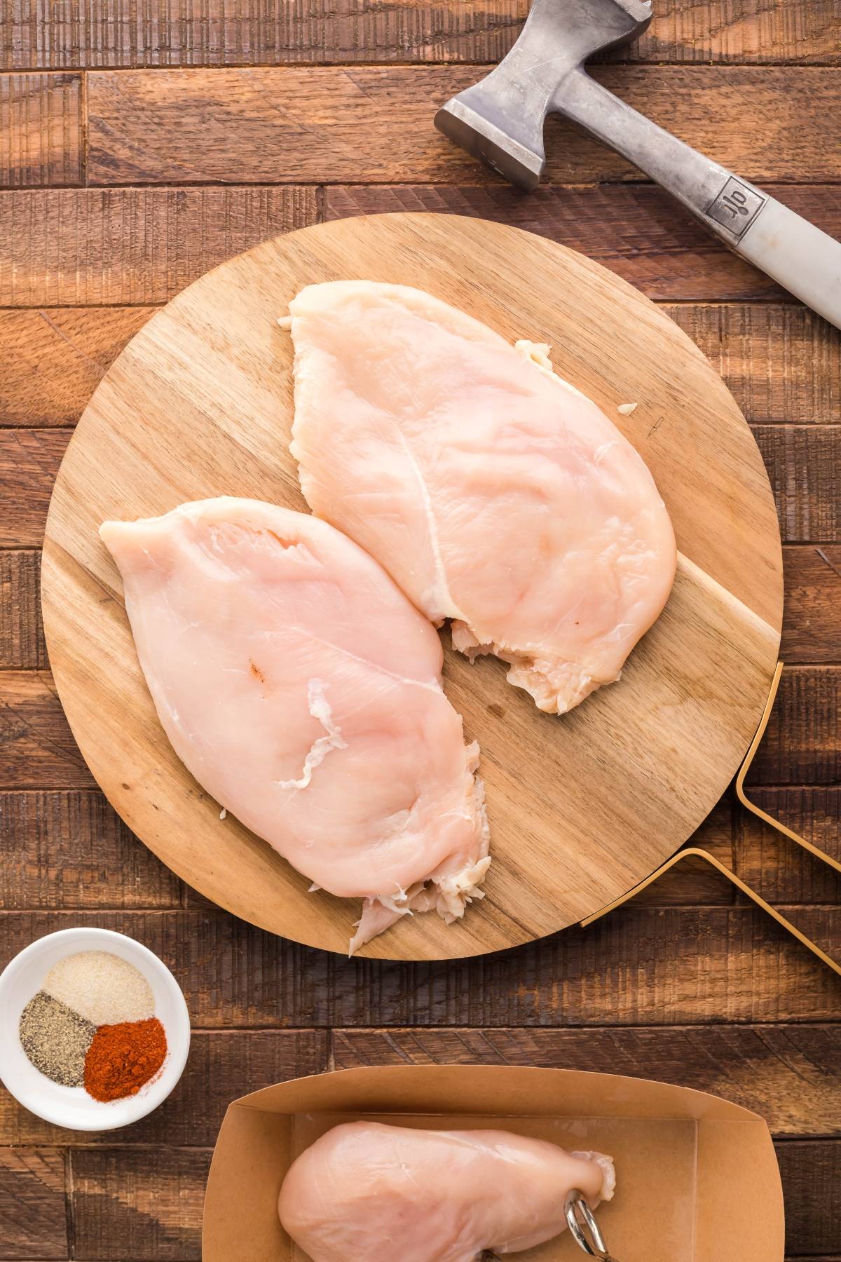 Two raw chicken breasts rest on a round wooden board with a meat mallet, seasonings, and another breast nearby—perfect for preparing a summer BBQ chicken salad or a simple radicchio salad.