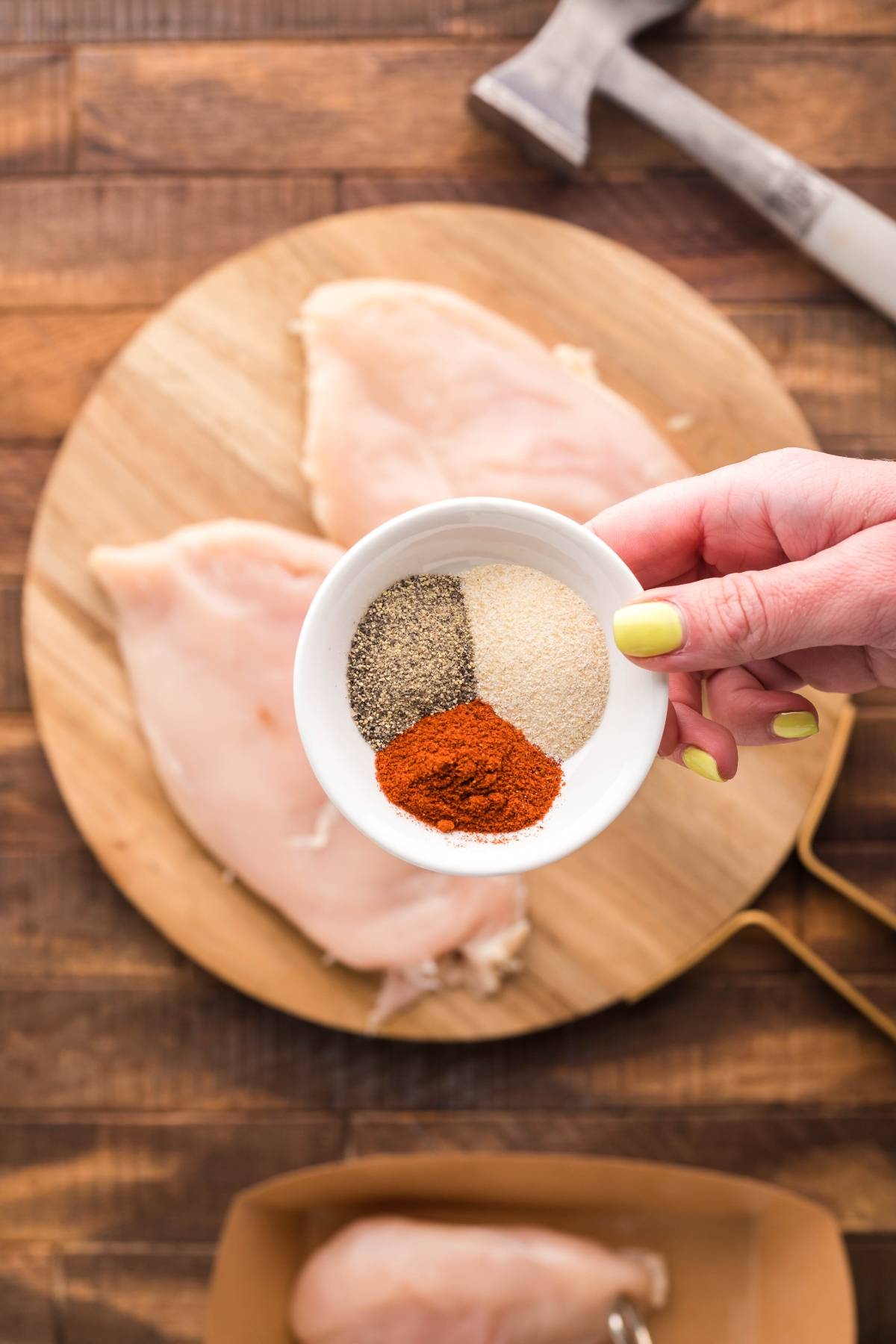 A hand holds a small bowl of spices above raw chicken breasts on a wooden cutting board, prepping flavorful chicken for a summer BBQ chicken salad.