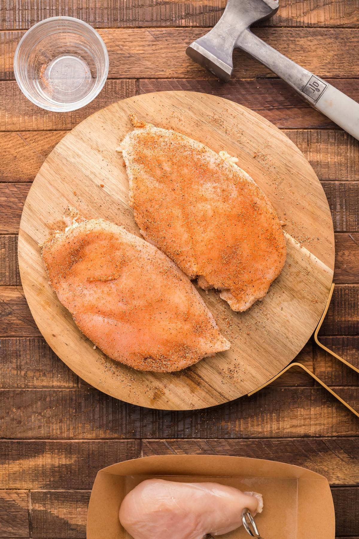 Two raw, seasoned chicken breasts on a wooden board, ready to be cooked for a Radicchio Endive Chicken Salad, with a meat mallet and glass of water nearby.