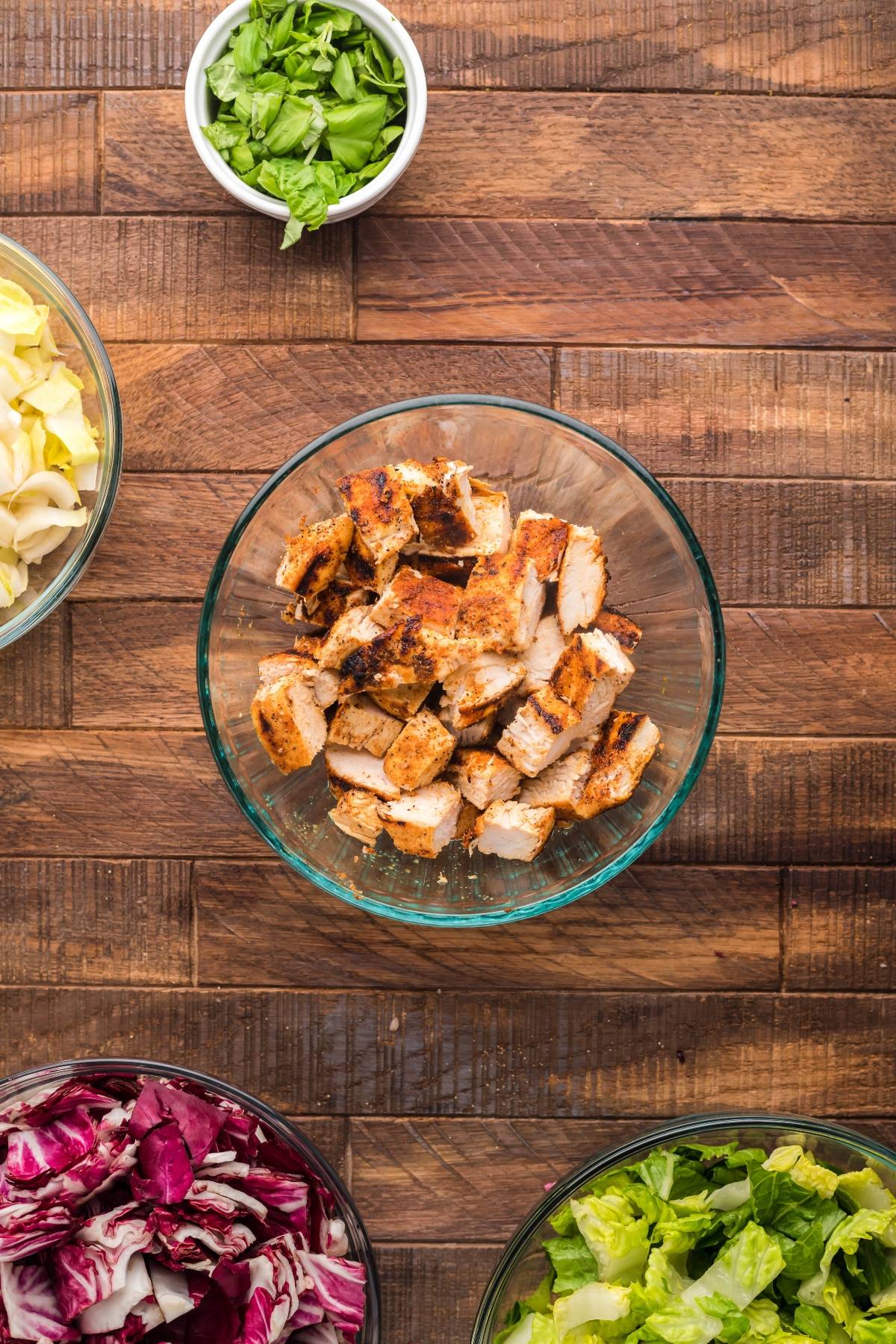 A bowl of chopped grilled chicken sits among bowls of greens and vegetables, ready to create a summer BBQ chicken salad with radicchio, endive, and a drizzle of creamy honey mustard dressing on a wooden table.