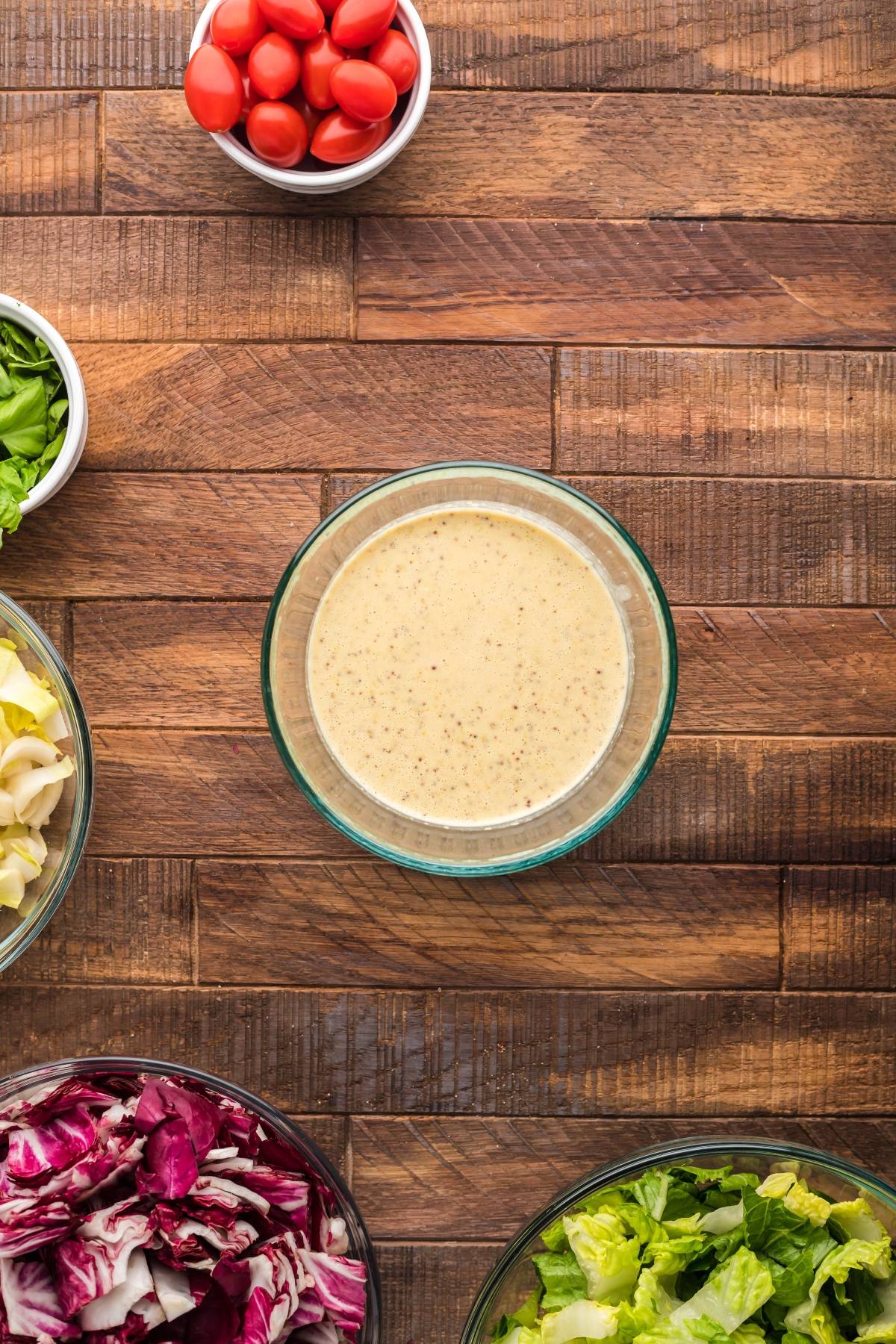 Glass bowl of Creamy Honey Mustard Dressing surrounded by lettuce, radicchio, tomatoes, and artichoke—perfect for a simple radicchio salad or to pair with your summer BBQ chicken salad on a wooden table.