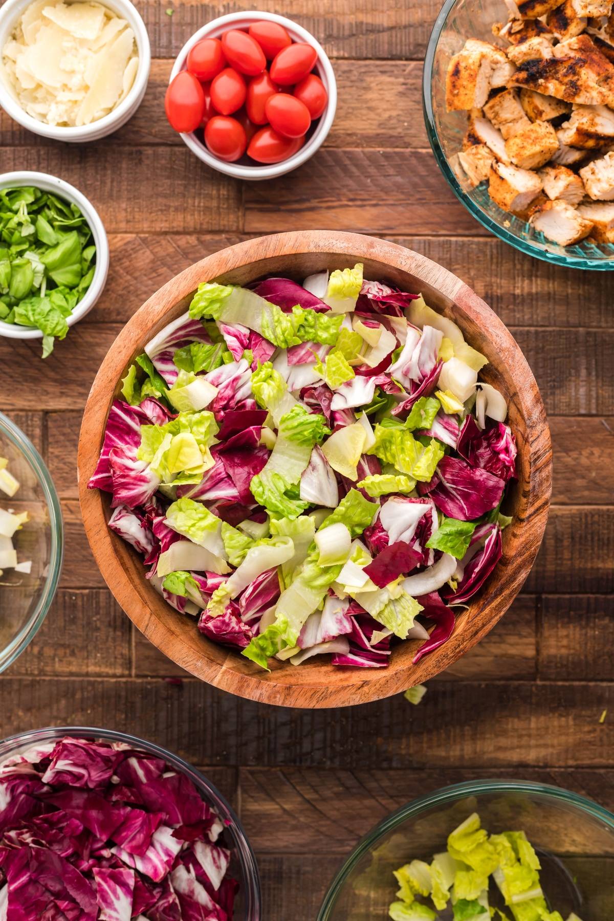 Wooden bowl of mixed salad greens, perfect for a simple radicchio salad, surrounded by small bowls of cherry tomatoes, chicken, cheese, herbs, and a Creamy Honey Mustard Dressing.