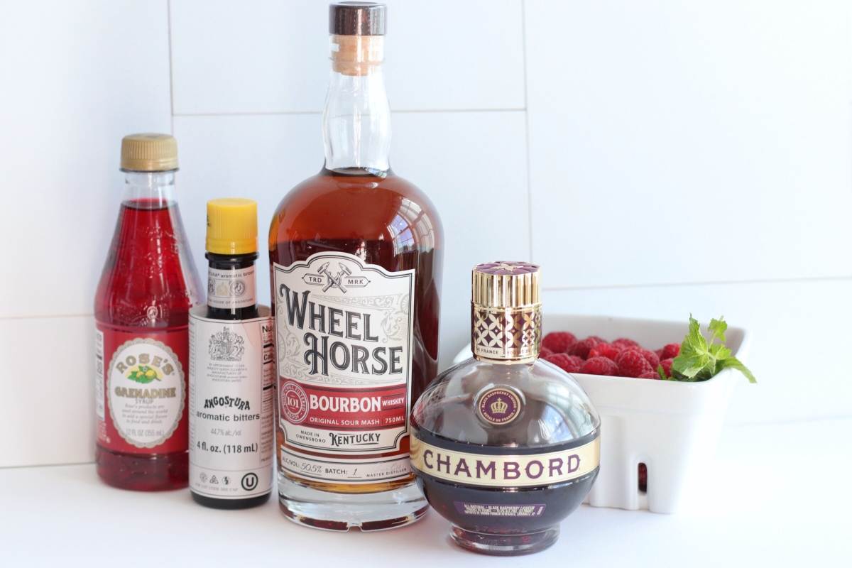 Bottles of bourbon, Chambord raspberry liqueur, grenadine, bitters, and a basket of fresh raspberries on a white surface—perfect for crafting a Raspberry Bourbon Smash Cocktail or any cocktail with raspberries.