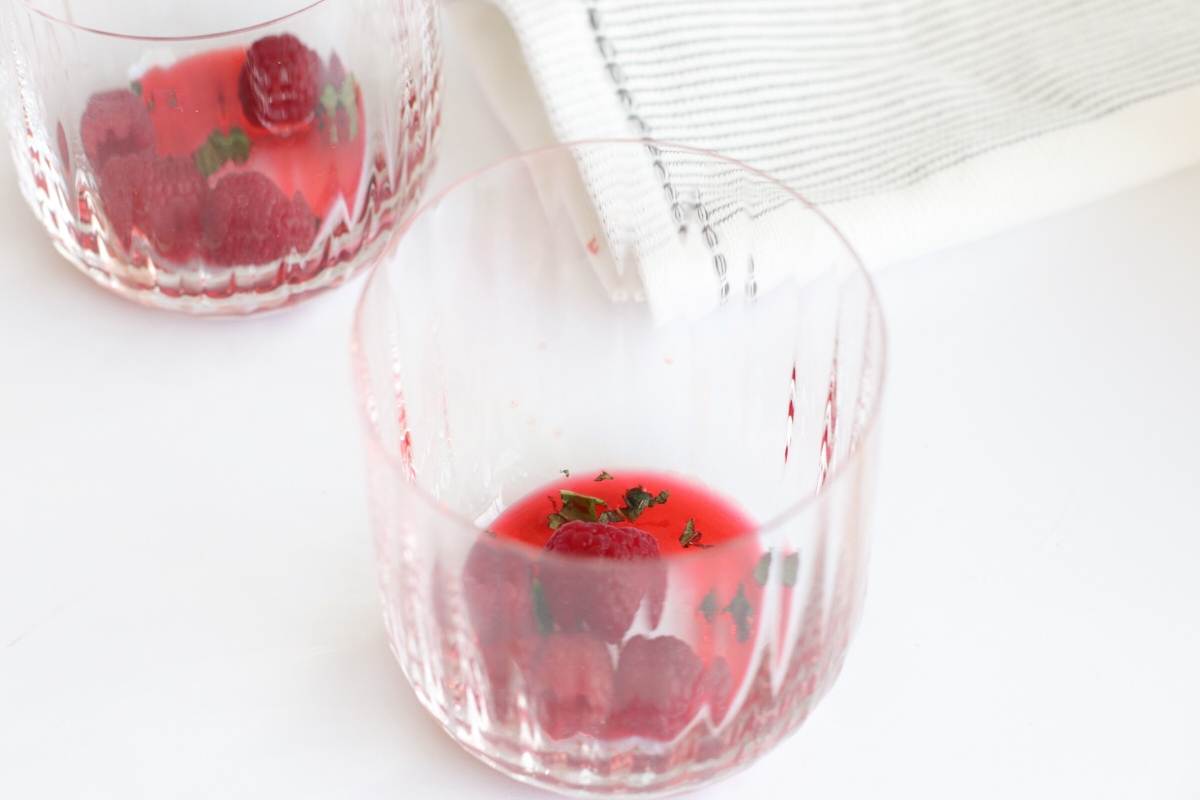 Two clear glasses with raspberries, juice, and herbs—perfect for trying a refreshing bourbon and Chambord raspberry liqueur cocktail—are set beside a folded white and gray striped cloth.