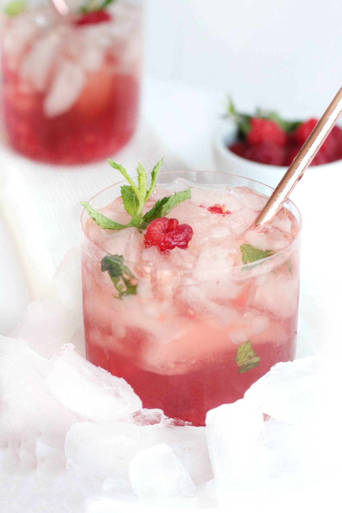 A glass of iced raspberry cocktail made with bourbon and Chambord raspberry liqueur, garnished with mint leaves and a straw, surrounded by ice cubes.