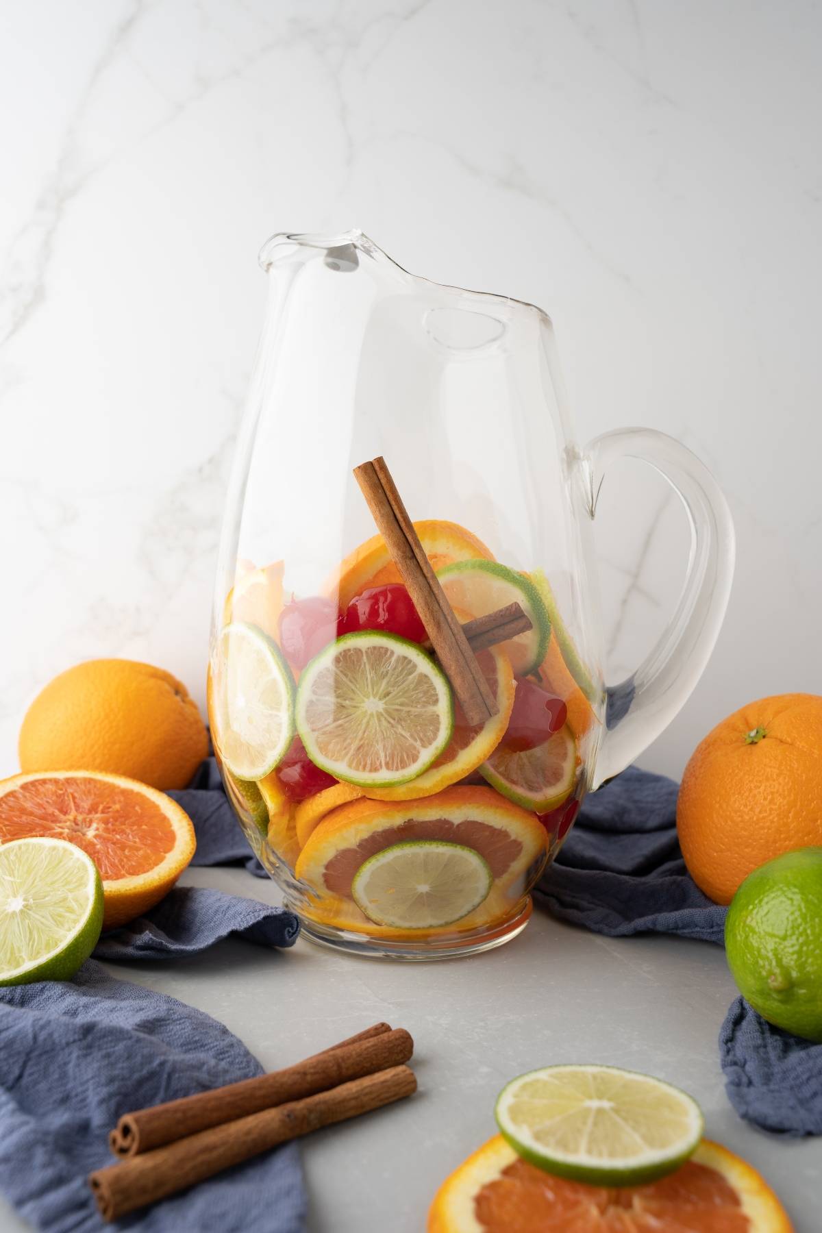 A glass pitcher filled with sliced citrus fruits and cinnamon sticks, surrounded by whole fruits, perfect for serving your favorite rum punch sangria at summer gatherings.