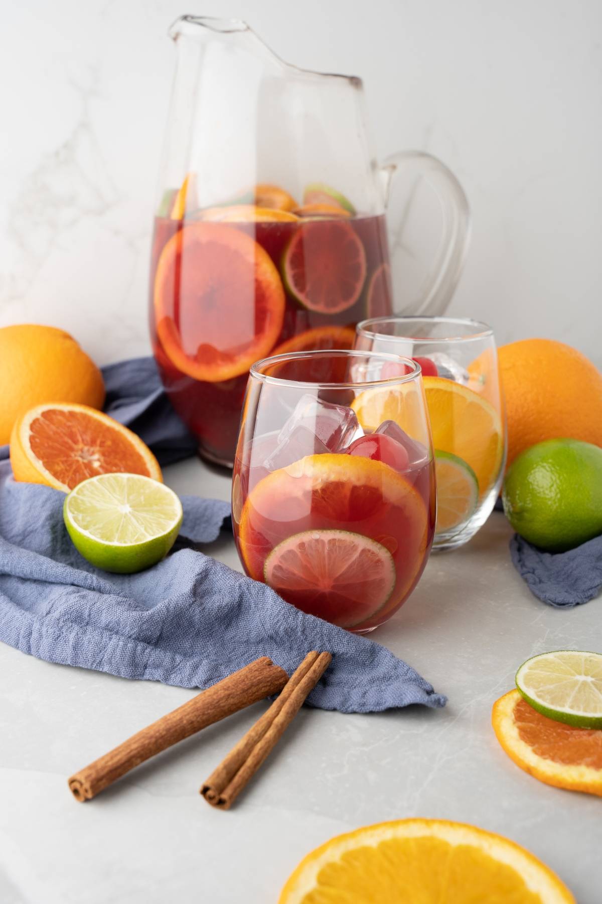 A glass and pitcher of rum punch sangria with orange, lemon, and lime slices, surrounded by fresh fruit and cinnamon sticks.
