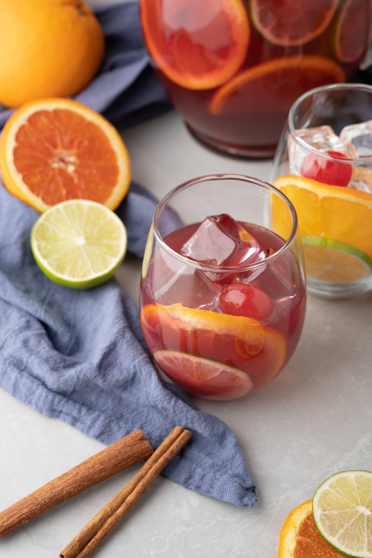 A glass of cocktail with red wine and rum, filled with ice, orange slices, and cherries, surrounded by fresh fruit and cinnamon sticks.