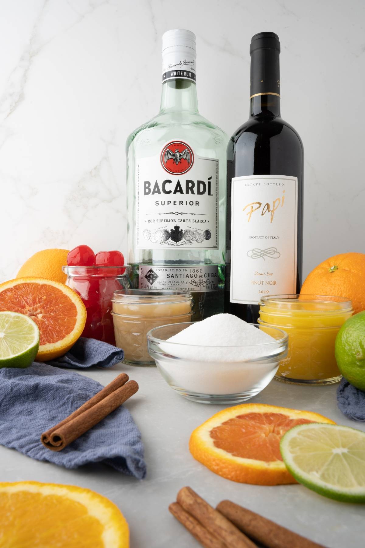 Bacardi rum, red wine, fruit, and cinnamon sticks on a marble counter with sugar and juice in bowls—perfect ingredients for the best summer sangria recipe or a flavorful cocktail with red wine and rum.