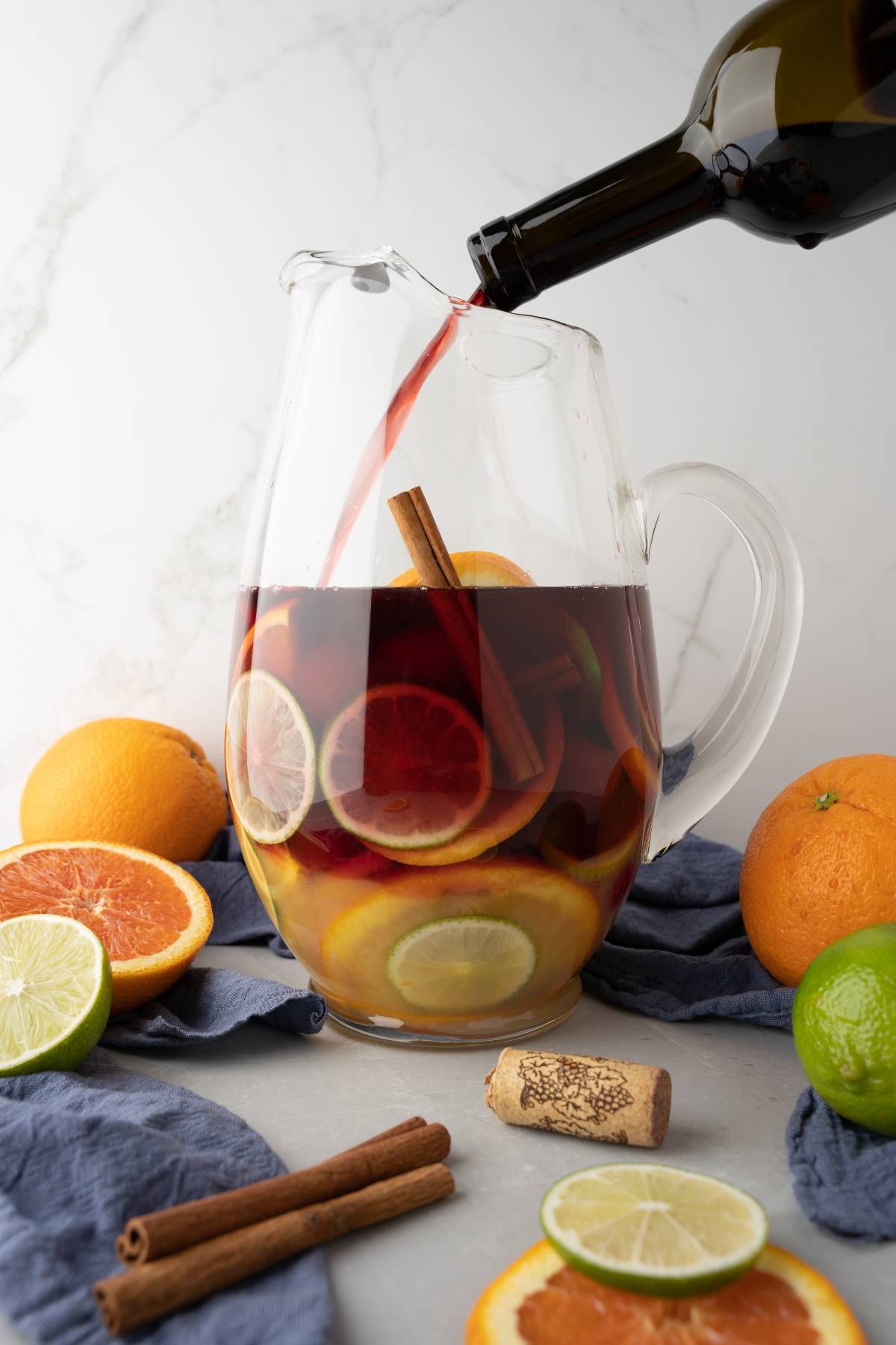Red wine is being poured into a glass pitcher with citrus slices and cinnamon sticks, surrounded by fruit, creating the best summer sangria recipe—an inviting cocktail with red wine and rum perfect for any gathering.