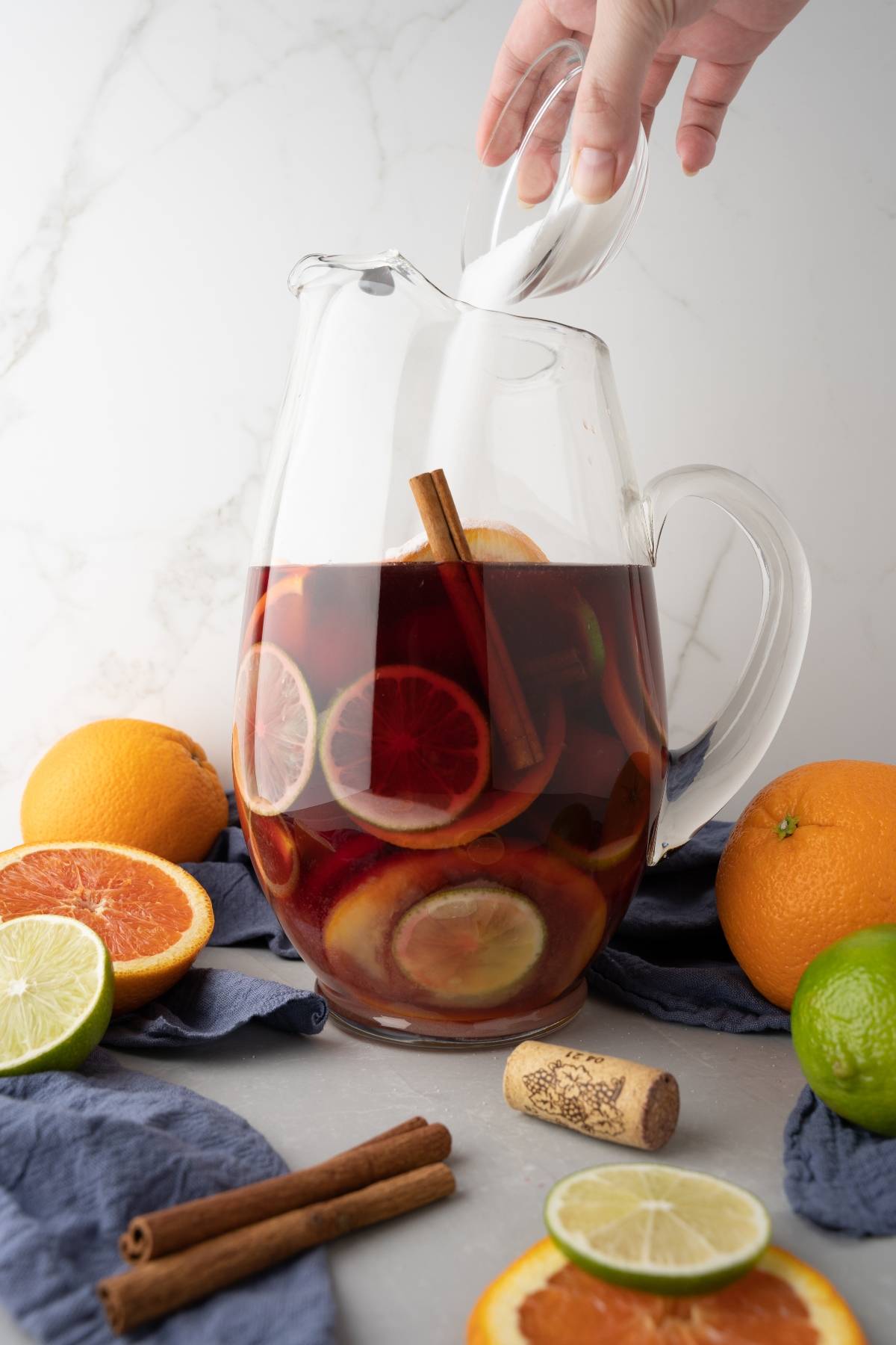 A hand pours sugar into a glass pitcher of rum punch sangria with citrus slices and cinnamon sticks, surrounded by fruit.
