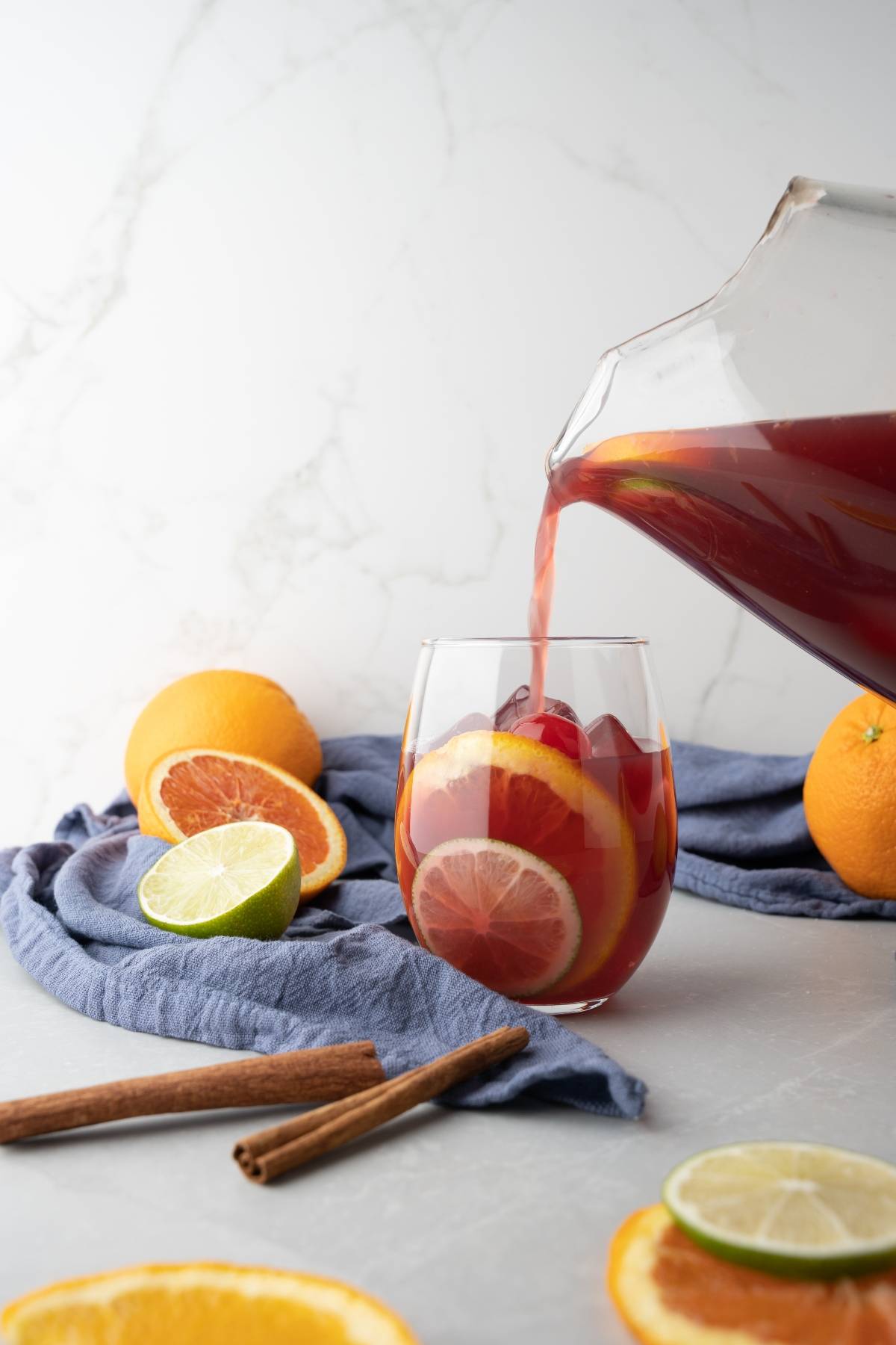 A pitcher pours red juice into a glass with citrus slices, surrounded by fruit and cinnamon sticks—the perfect setup for the best summer sangria recipe.