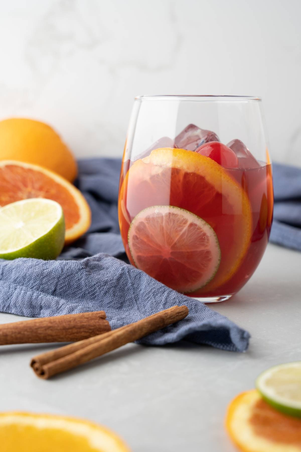 A glass of sangria with ice and citrus slices, surrounded by fruit and cinnamon sticks on a gray surface—an inviting cocktail with red wine and rum, perfect for summer gatherings.