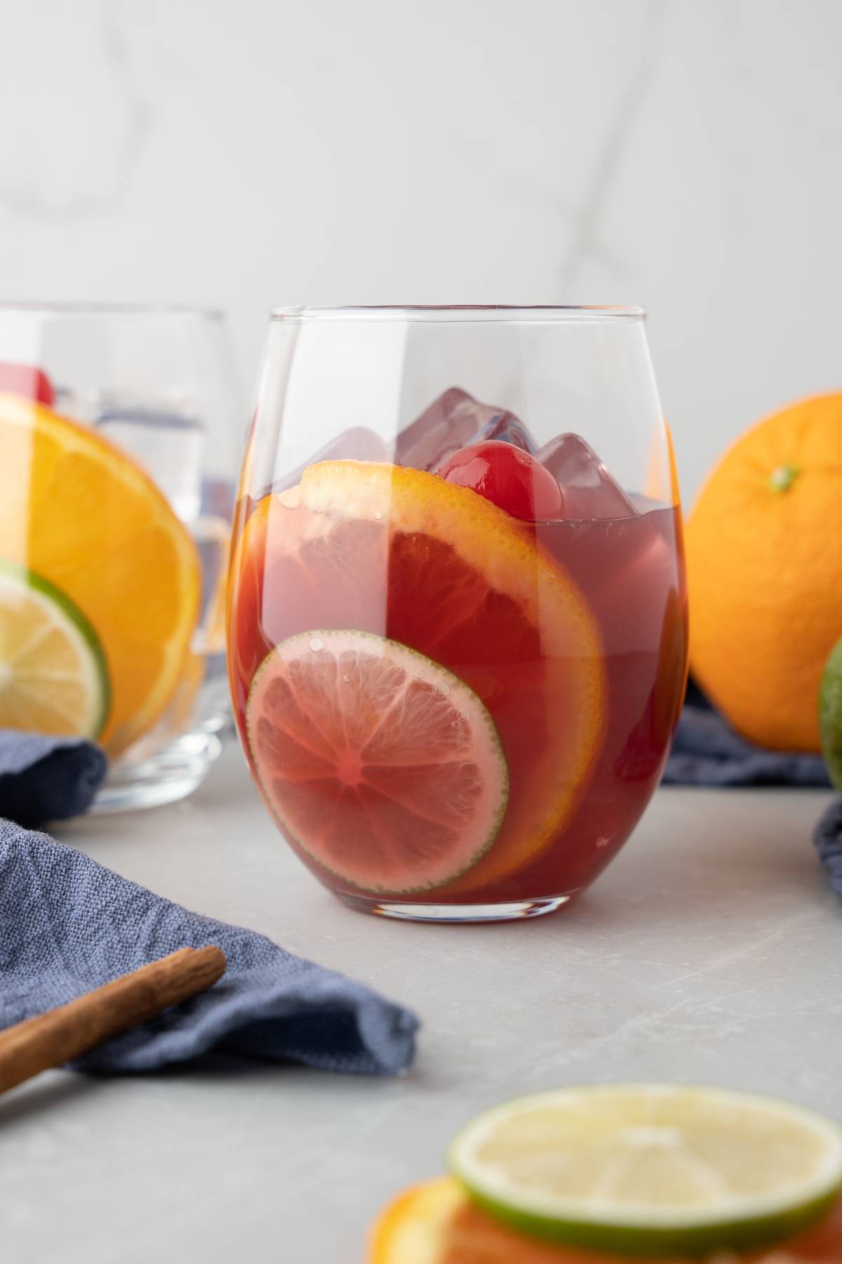 A glass of rum punch sangria with ice, orange, and lime slices sits on a gray table with fruit and a blue napkin nearby.