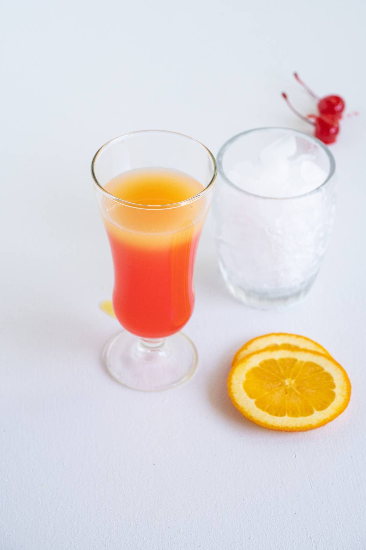 A glass of orange-red juice beside ice in a glass, orange slices, and two cherries on a white surface—perfect inspiration for your next Caribbean punch cocktail recipe.