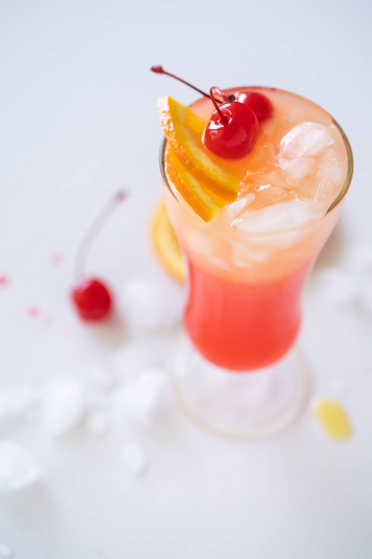 A tropical cocktail with ice, orange slices, and cherries, garnished with a cherry, on a white surface—an easy rum runner recipe perfect for summer sipping.