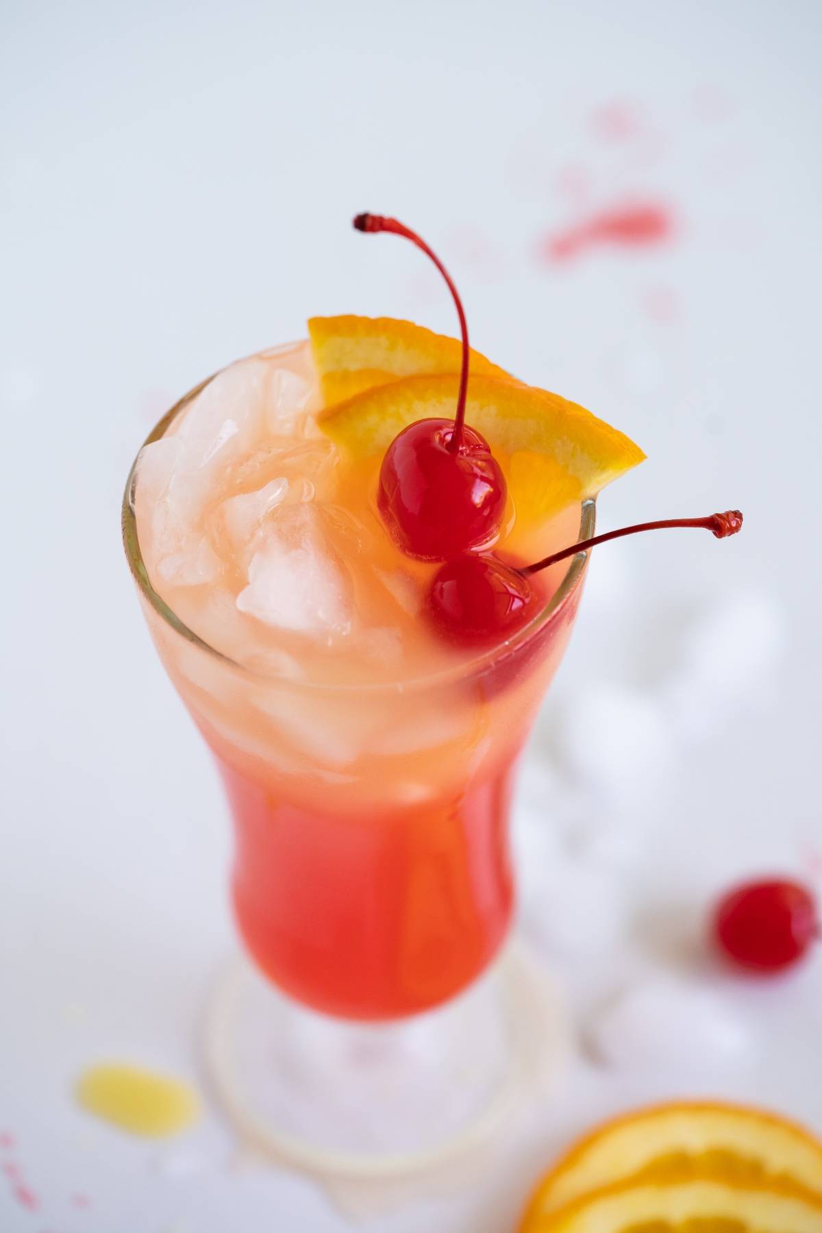 A tropical cocktail with ice, orange slice, and cherries garnish in a tall glass on a white surface—perfect for trying a refreshing Caribbean punch cocktail recipe.