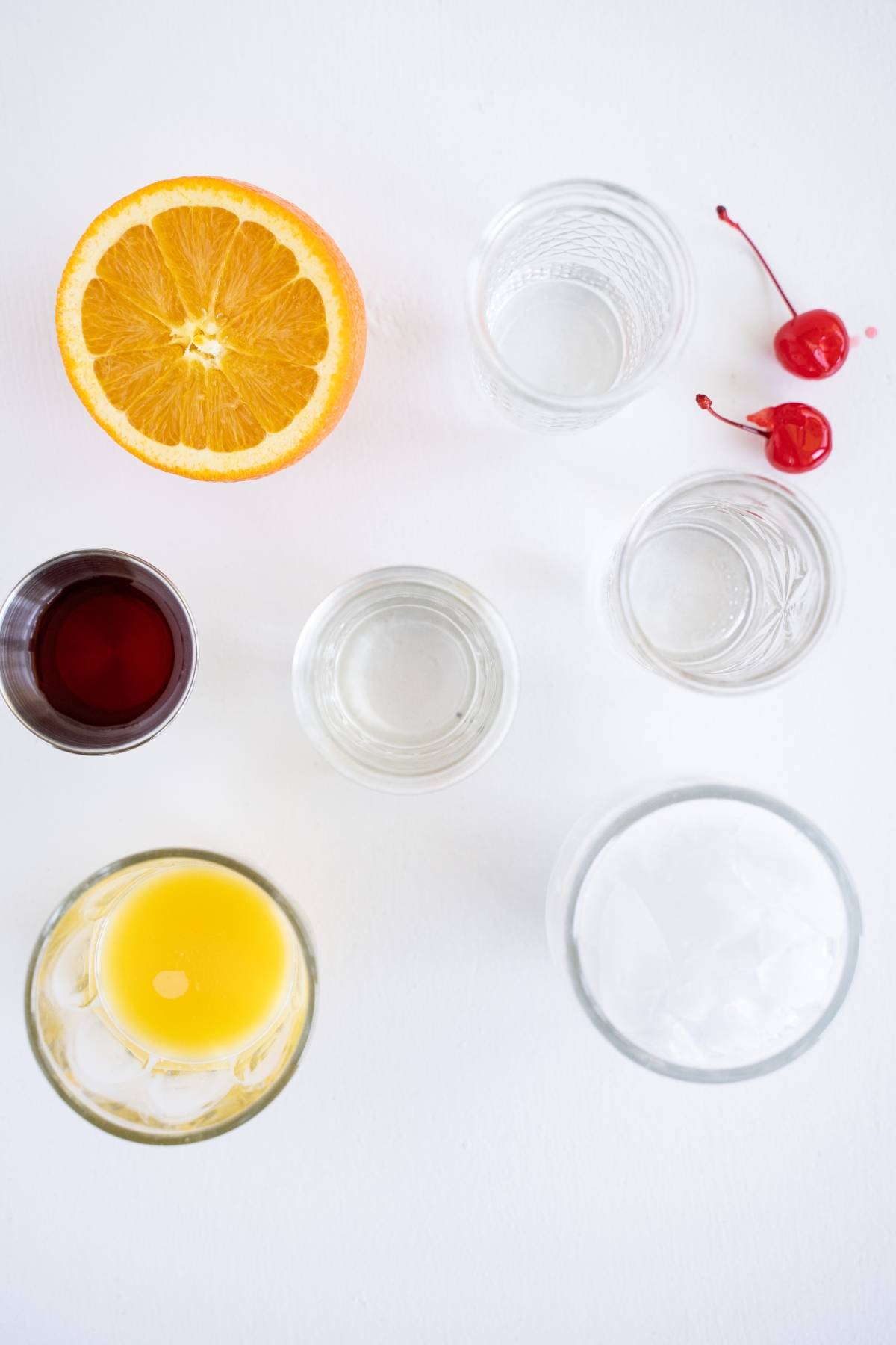 Orange slice, two cherries, and glasses of juice, liquor, and ice arranged on a white surface—perfect inspiration for an easy rum runner recipe.