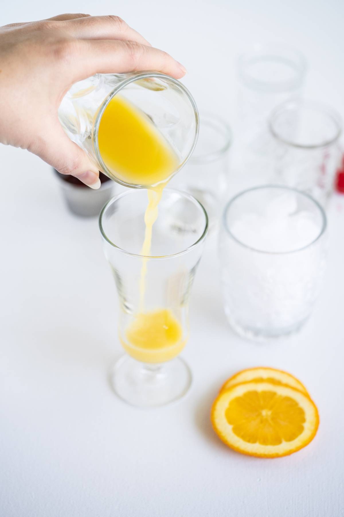 A hand pours orange juice into a glass beside ice-filled glasses and orange slices, creating the perfect base for a Pineapple rum drink recipe or a refreshing rum runner cocktail on a white surface.