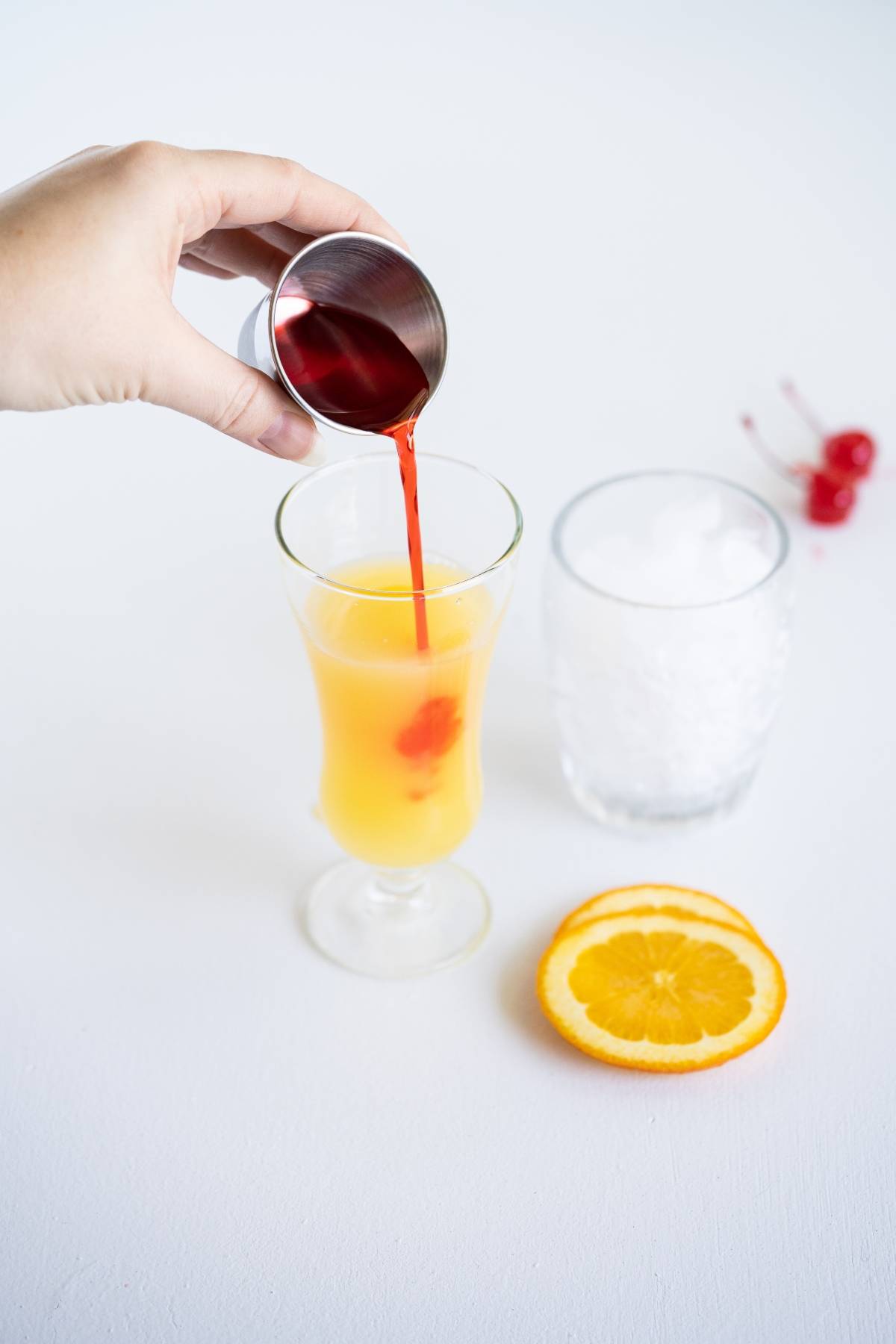 Hand pouring red syrup into an orange drink with ice, orange slices, and cherries on a white surface—perfect inspiration if you're searching how to make a rum runner or want a colorful Caribbean punch cocktail recipe.