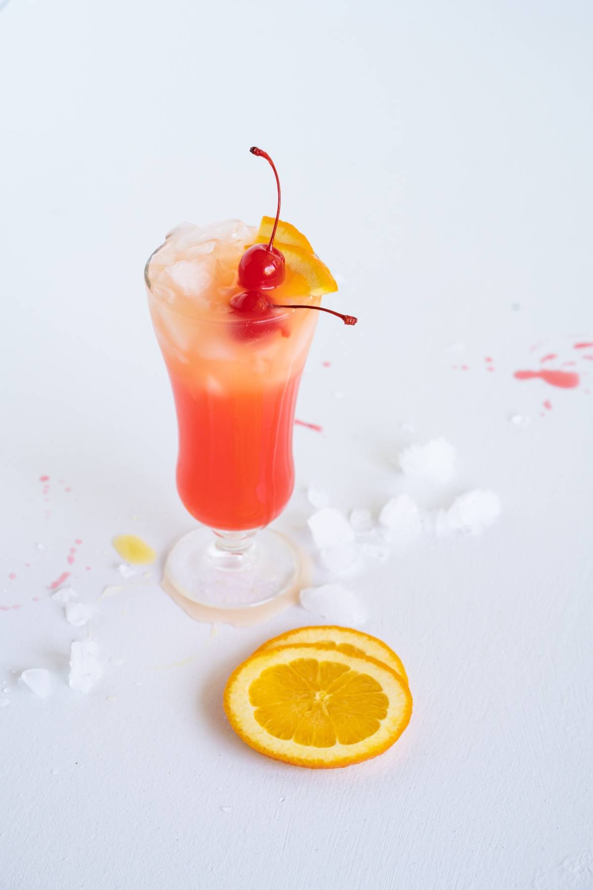 A colorful cocktail with ice, orange slice, and cherries, next to two orange slices on a white surface—perfect inspiration for your next Caribbean punch cocktail recipe.