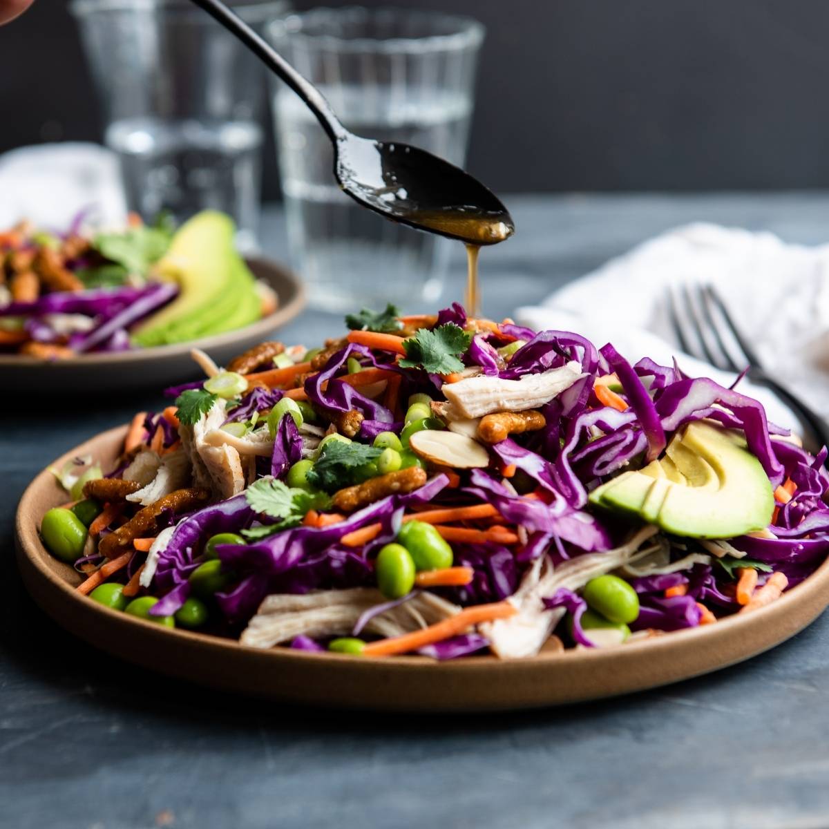 A plate of colorful salad with avocado, cabbage, edamame, and carrots is being drizzled with a vibrant honey ginger dressing—a delicious twist on the classic cabbage salad recipe.