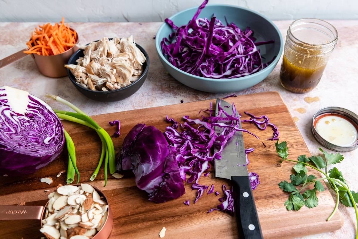 Chopped purple cabbage, shredded carrots, chicken, sauce, herbs, and creamy avocado come together for a fresh Cabbage salad recipe on a cutting board and table.