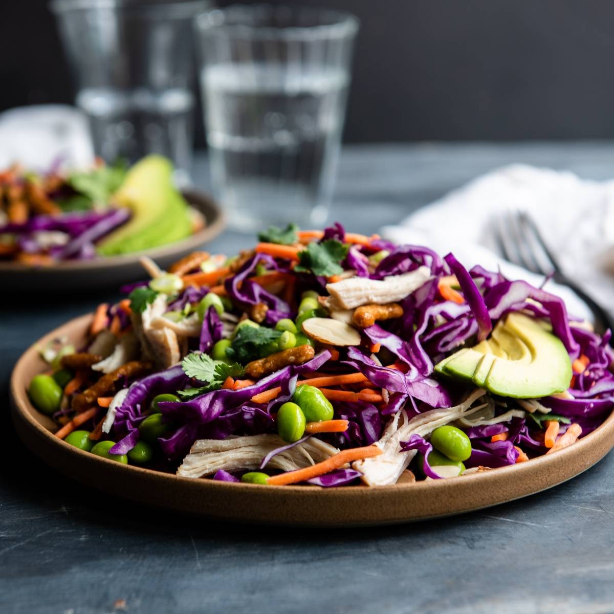 A vibrant cabbage salad recipe featuring purple cabbage, carrots, edamame, avocado, herbs, and nuts on a brown plate—delicious with a drizzle of honey ginger dressing.