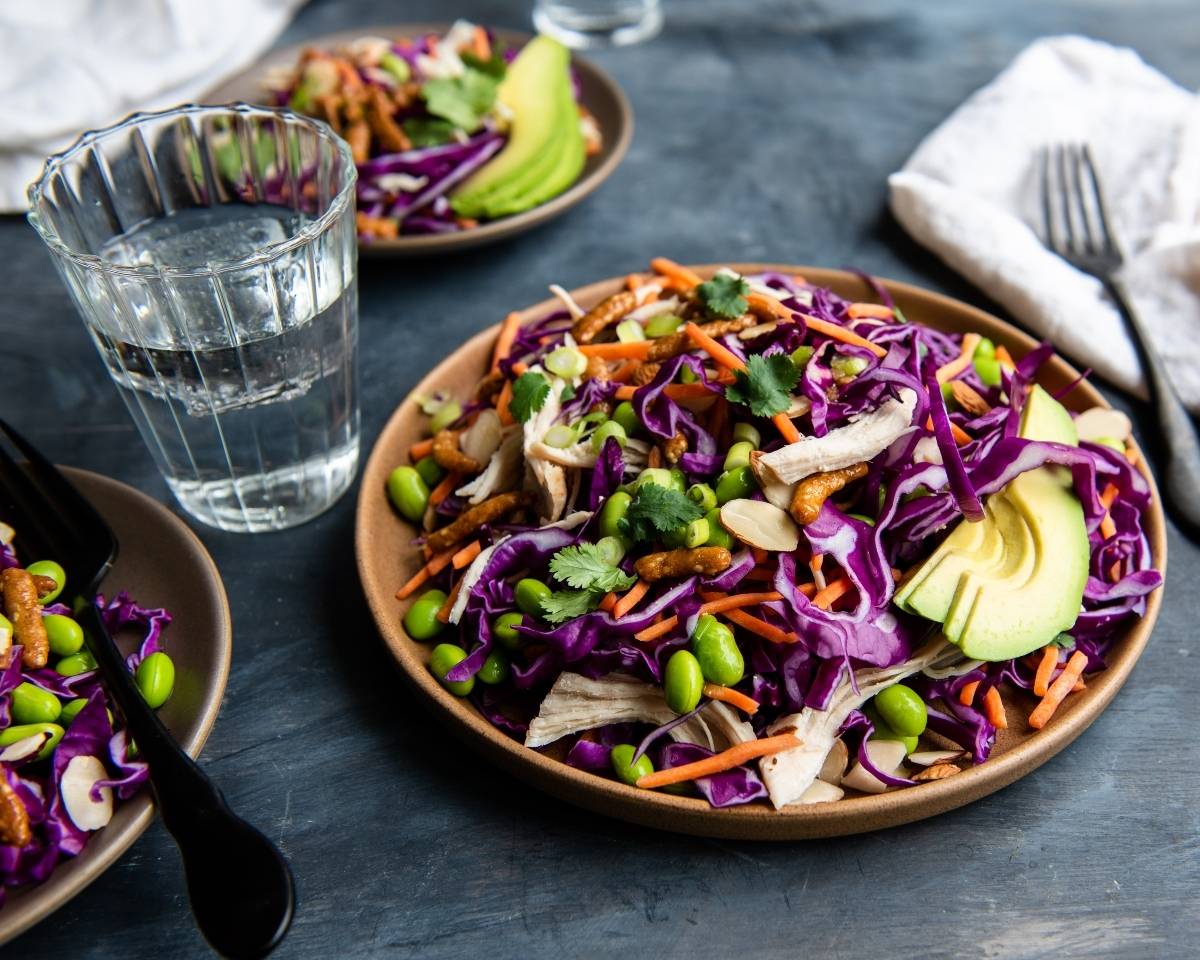 This vibrant Sesame Avocado Asian Chicken Salad features purple cabbage, avocado, edamame, shredded chicken, carrots, and fresh herbs on a brown plate—a tasty twist on a classic cabbage salad recipe.