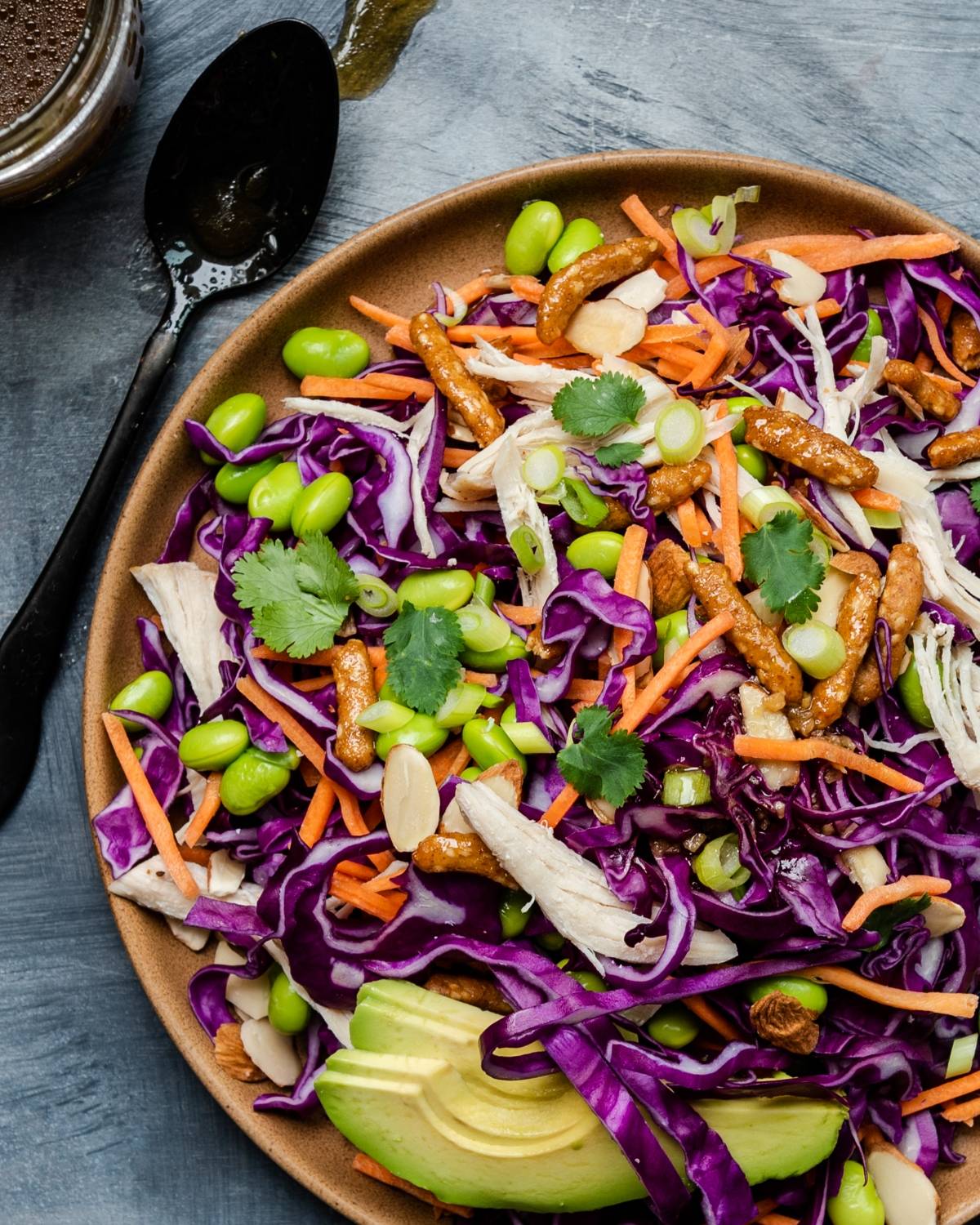 Colorful salad with red cabbage, carrots, avocado, edamame, cilantro, nuts, and crunchy toppings on a brown plate—served with a zesty honey ginger dressing for the perfect salad with avocado.