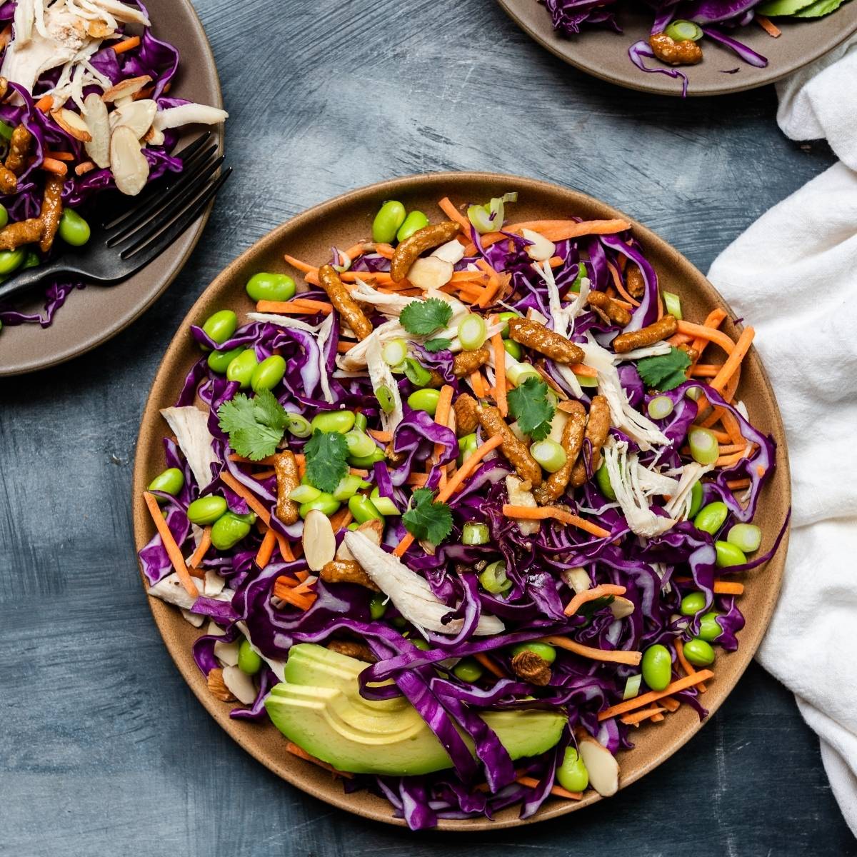 Colorful Sesame Avocado Asian Chicken Salad with purple cabbage, carrots, edamame, cilantro, and chicken on a brown plate—perfect with a drizzle of honey ginger dressing. Try this vibrant cabbage salad recipe for a fresh meal!.