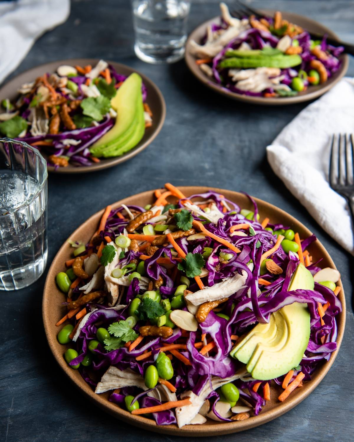 Three plates of colorful cabbage chicken salad with avocado slices, carrots, edamame, almonds, and cilantro are served next to glasses of water—deliciously paired with a honey ginger dressing.