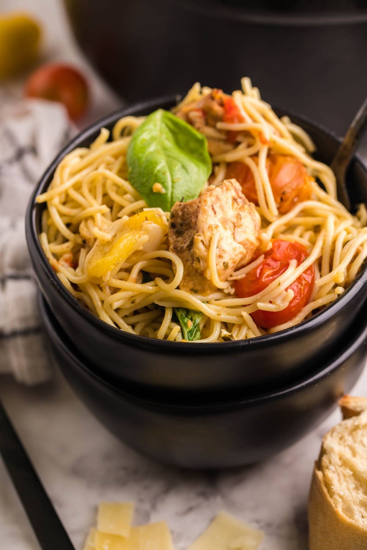 A black bowl of baked chicken caprese with pasta featuring chicken tenderloins, cherry tomatoes, and basil, served with bread and cheese on the side.