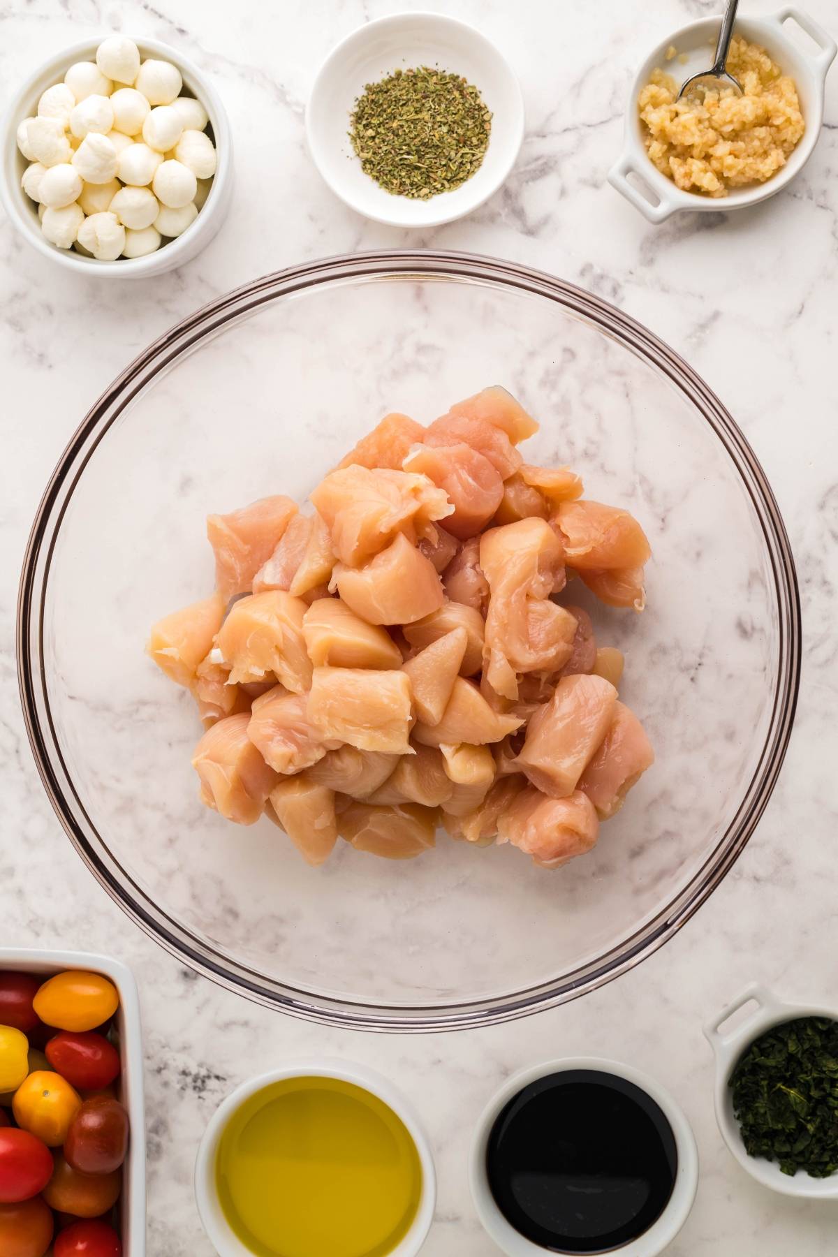 A bowl of raw diced chicken tenderloins is surrounded by seasonings, oil, tomatoes, mozzarella balls, and garlic on a marble counter—perfect ingredients for an easy chicken dinner or easy oven chicken caprese.