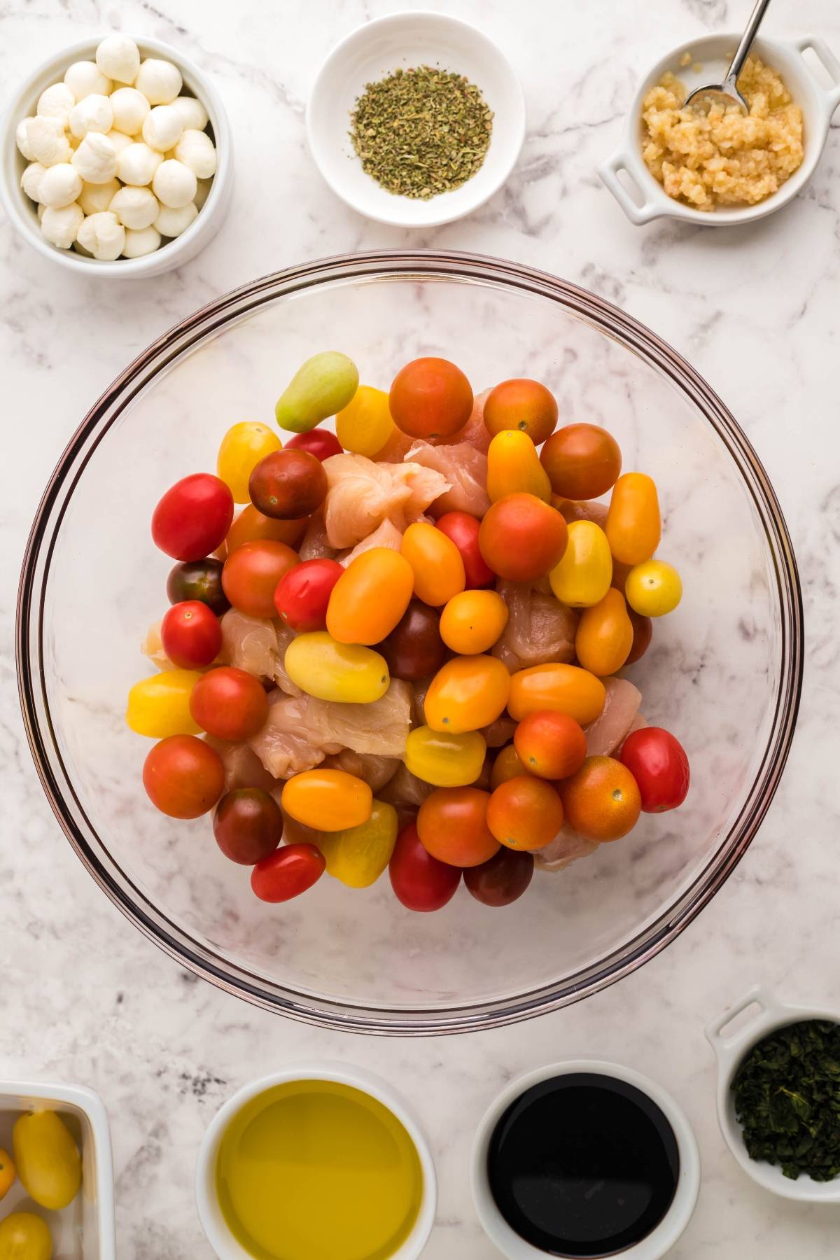 A bowl of cherry tomatoes and chicken, inspired by easy oven chicken caprese, is surrounded by small bowls of seasonings, oil, vinegar, and cheese.