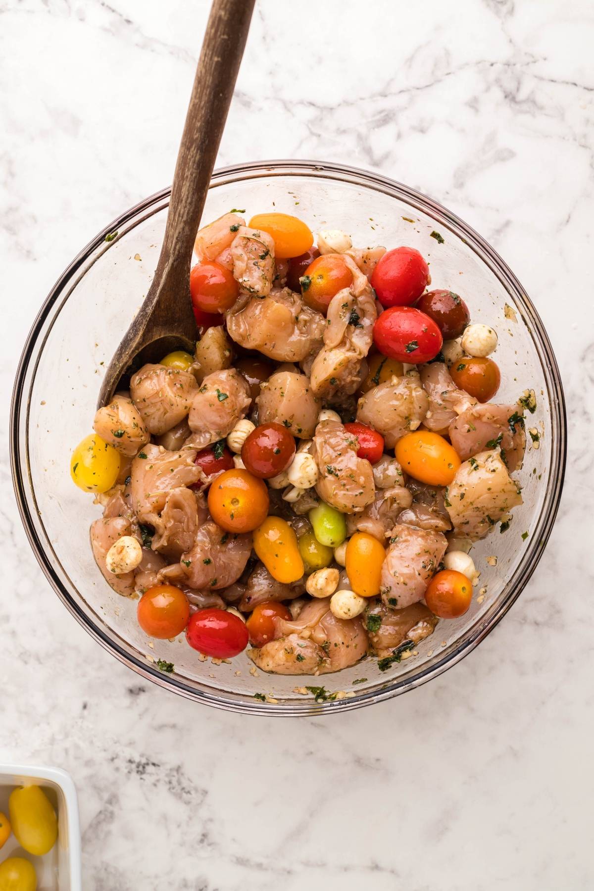 A glass bowl of marinated chicken tenderloins, cherry tomatoes, and herbs with a wooden spoon on a marble surface—perfect for an easy oven chicken caprese.