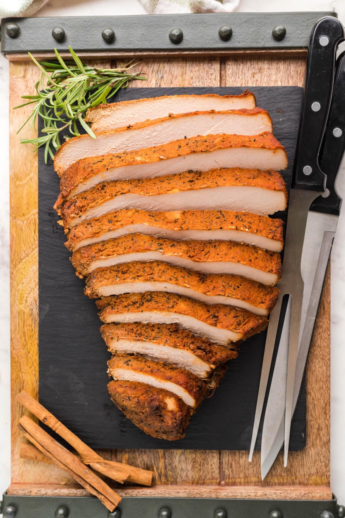 Sliced Slow Cooker Apple Cider Turkey Breast on a cutting board with herbs, cinnamon sticks, knife, and carving fork.