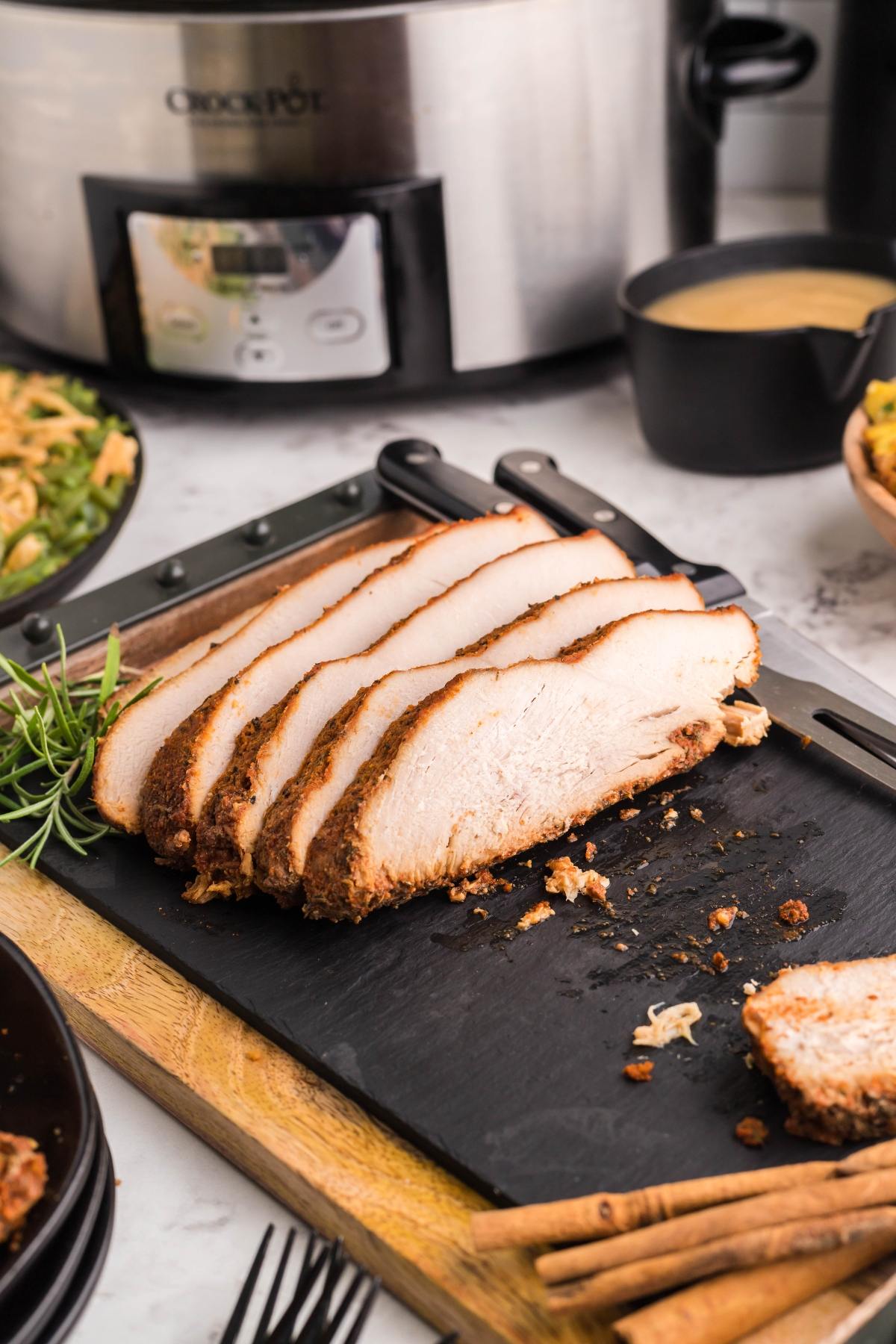 Sliced turkey breast on a black cutting board with herbs, beside a slow cooker and a bowl of gravy—perfect for trying our Slow Cooker Apple Cider Turkey Breast.
