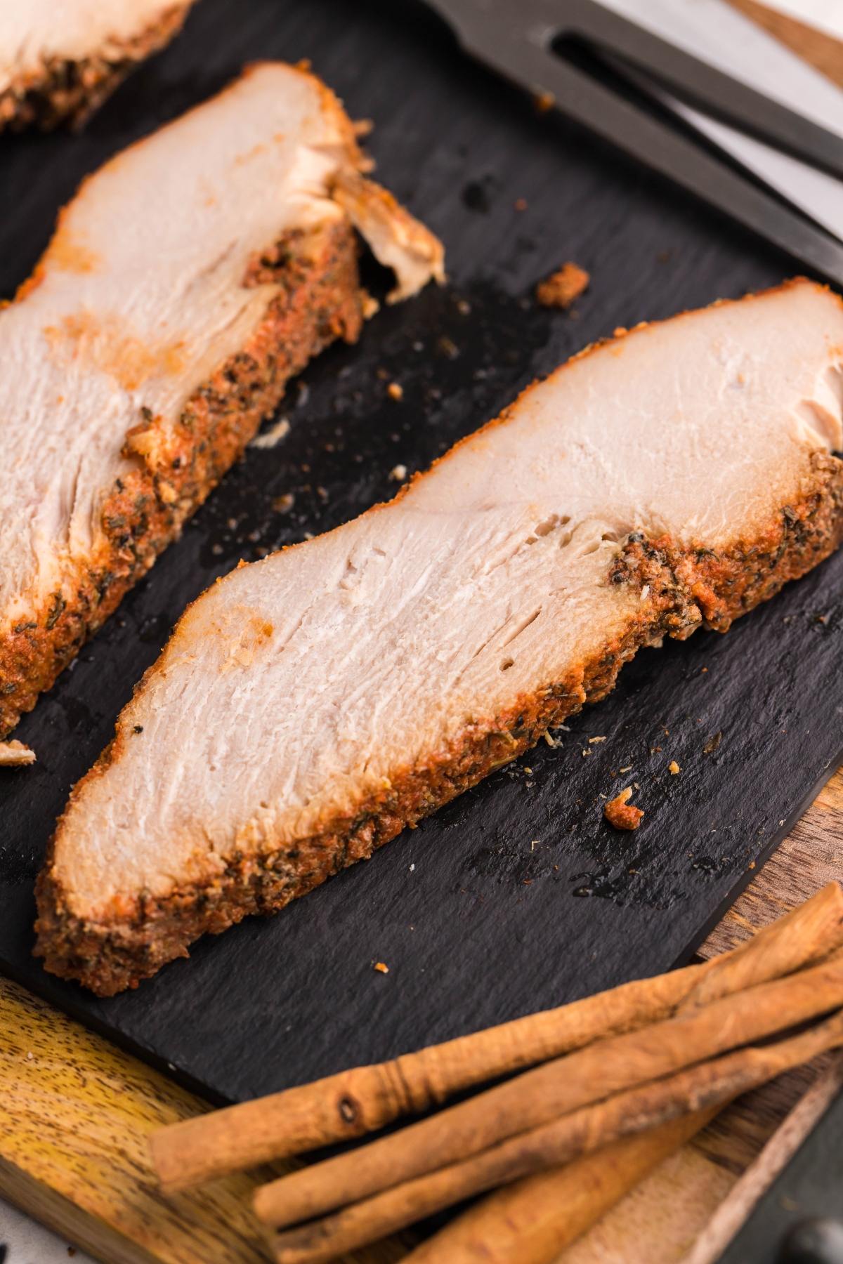 Two thick slices of seasoned turkey breast on a black slate with cinnamon sticks nearby, inspired by a Slow Cooker Apple Cider Turkey Breast recipe.