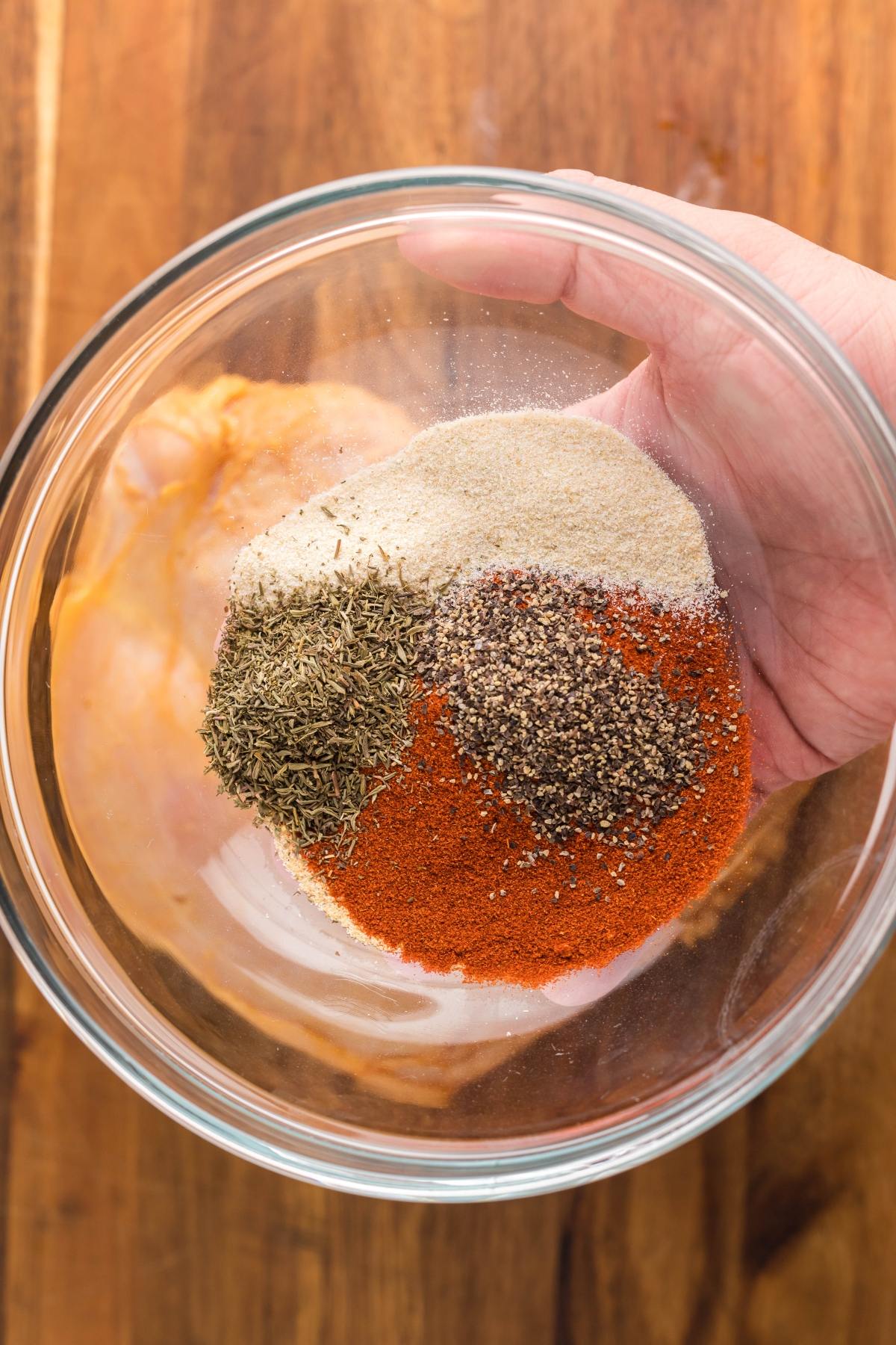 A hand holding a glass bowl with various colorful spices on top of raw chicken on a wooden surface, perfect for prepping an easy brine for turkey breast or starting your favorite slow cooker turkey breast recipe.