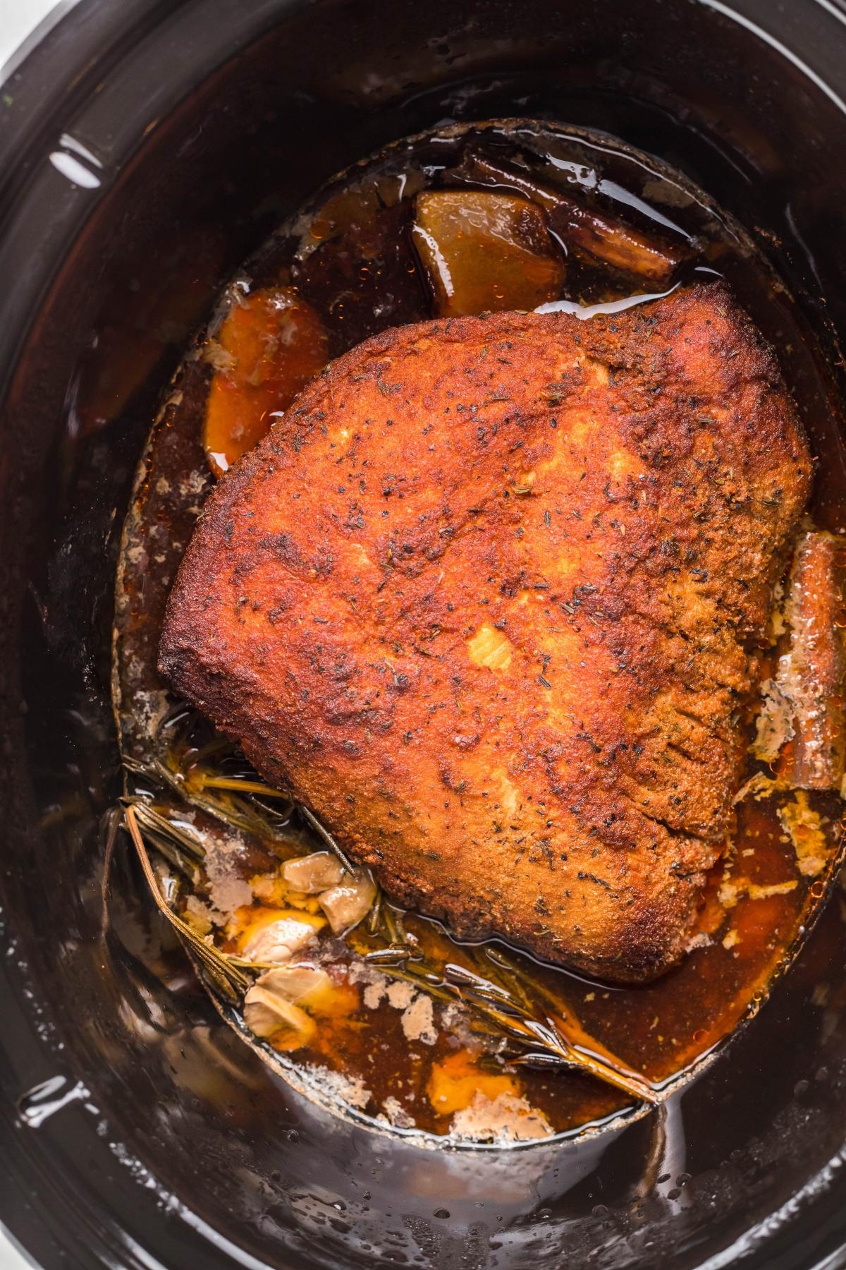 Seasoned pork roast cooking in a slow cooker with herbs and juices visible around the meat, much like preparing a boneless turkey breast in a slow cooker for tender, flavorful results.
