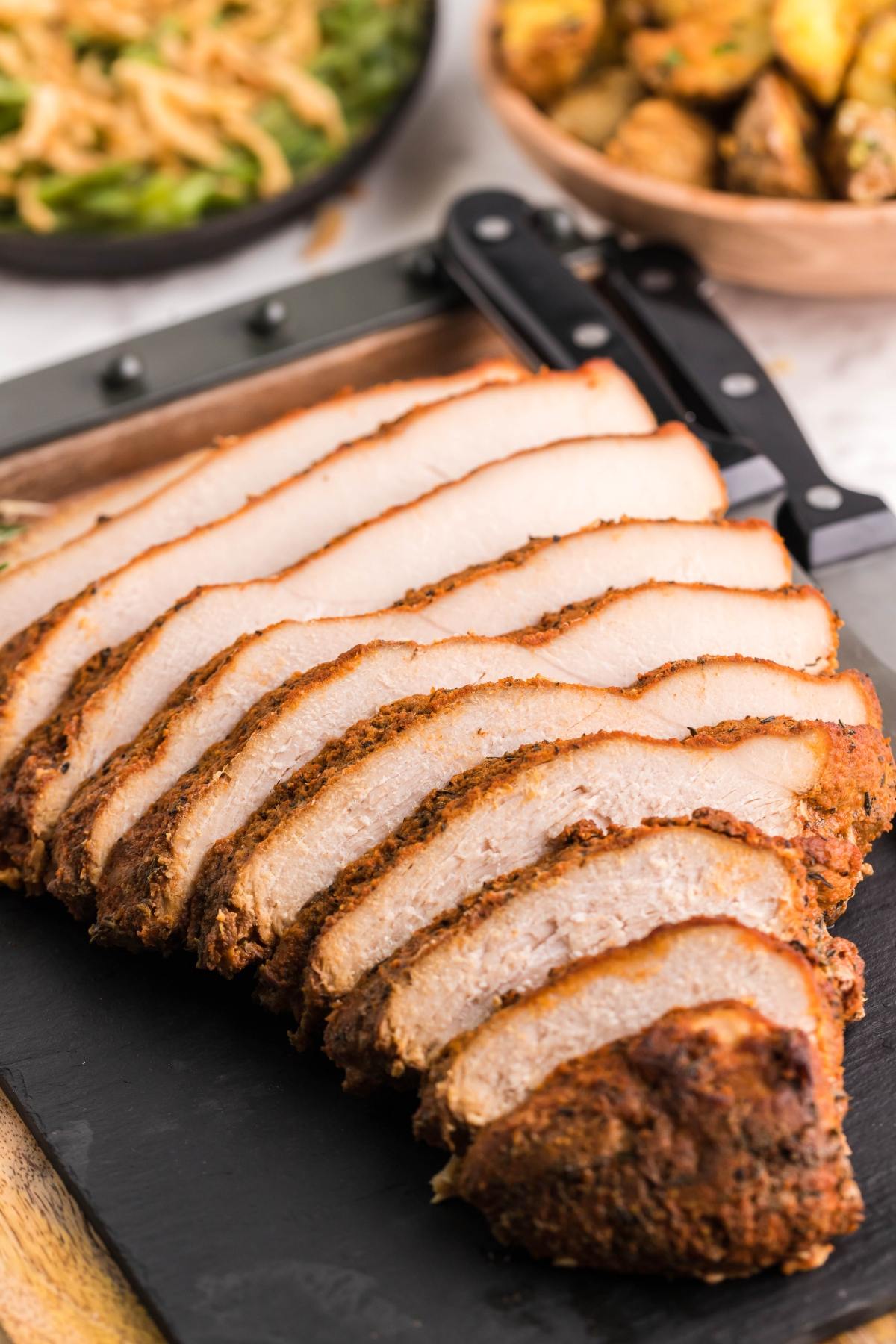 Sliced roast pork arranged on a black cutting board, with sides of green beans and roasted potatoes in the background—perfect inspiration for your next boneless turkey breast in a slow cooker.