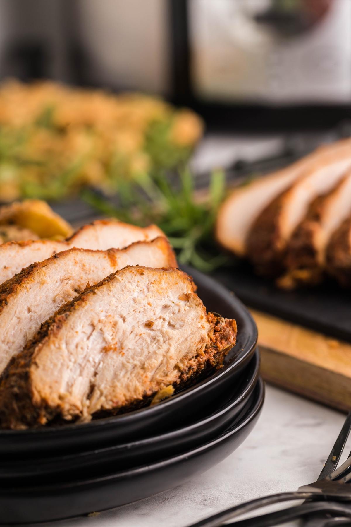 Close-up of sliced, seasoned pork on a black plate with more pork and greens blurred in the background, perfect for those who enjoy dishes like boneless turkey breast in a slow cooker.