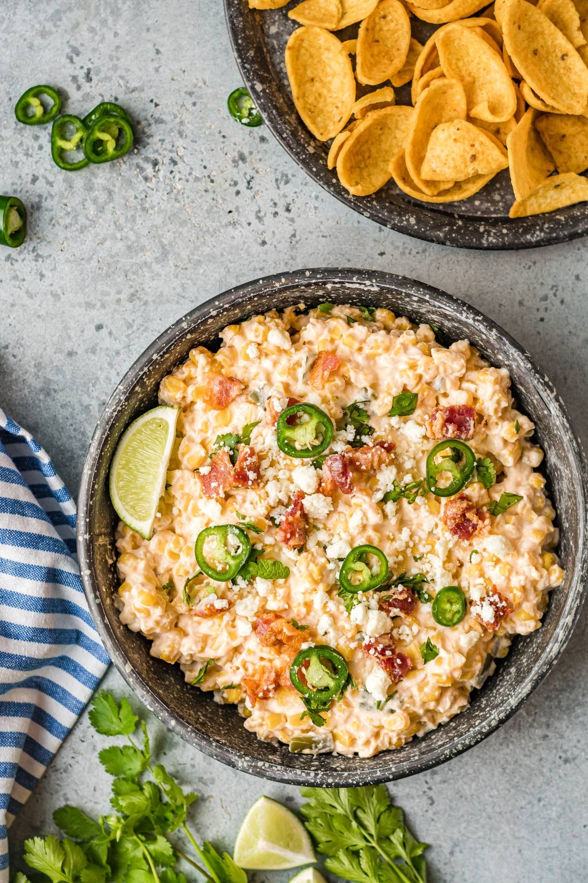 A bowl of creamy jalapeño corn dip topped with cheese, bacon, and a lime wedge sits next to crunchy corn chips—an irresistible, cheesy Mexican-style appetizer perfect for sharing.