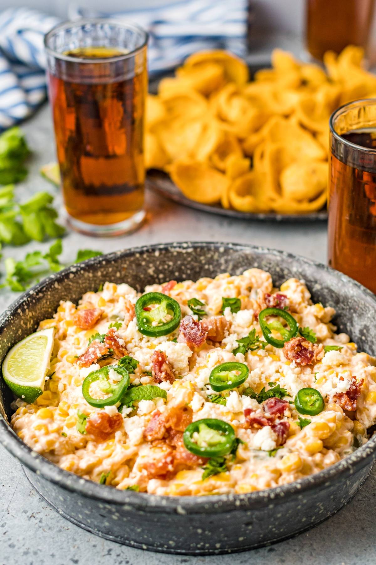A bowl of creamy jalapeño corn dip topped with bacon, cheese, and a lime wedge, served as a cheesy Mexican-style appetizer with drinks in the background.