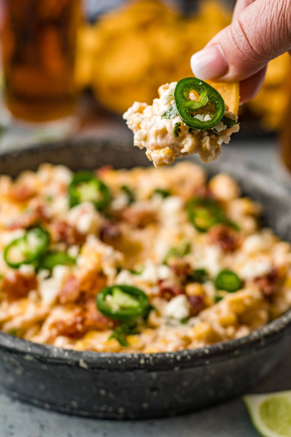 A hand holds a chip topped with cheesy Slow Cooker Jalapeño Corn Dip and sliced jalapeños above a bowl of the same irresistible slow cooker dip recipe.