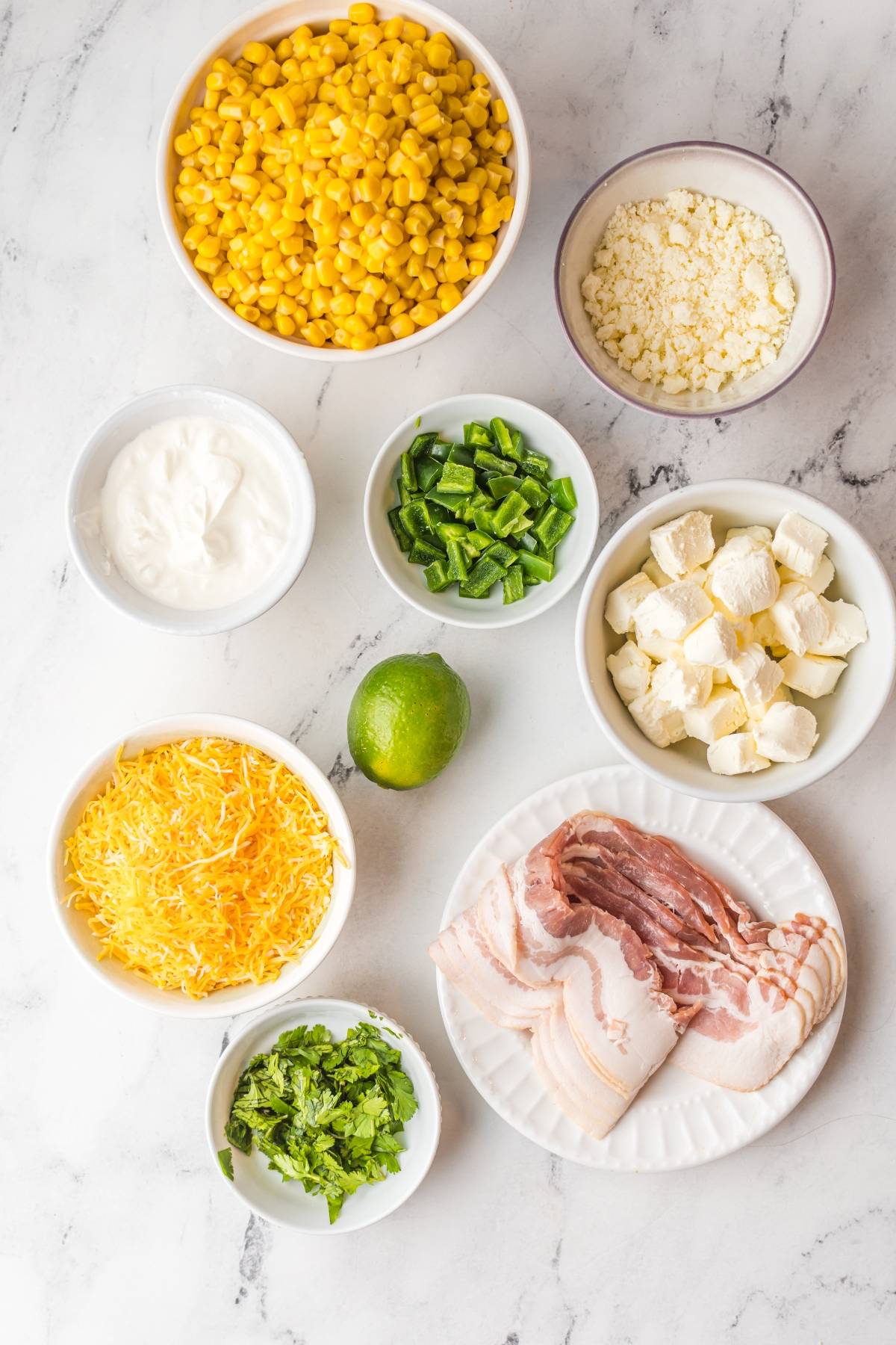 Ingredients laid out on a marble surface for an easy street corn dip: corn, cheese, bacon, lime, peppers, sour cream, and fresh herbs.